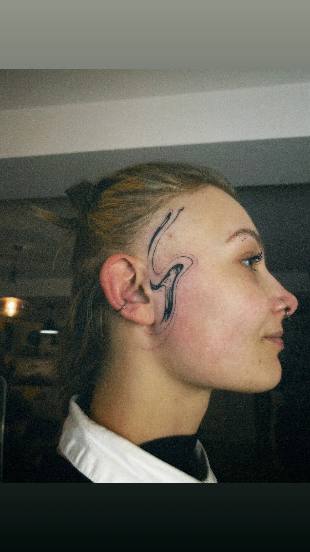 Side profile of a young woman with a facial tattoo on her temple and cheek, and multiple earrings in her ear.