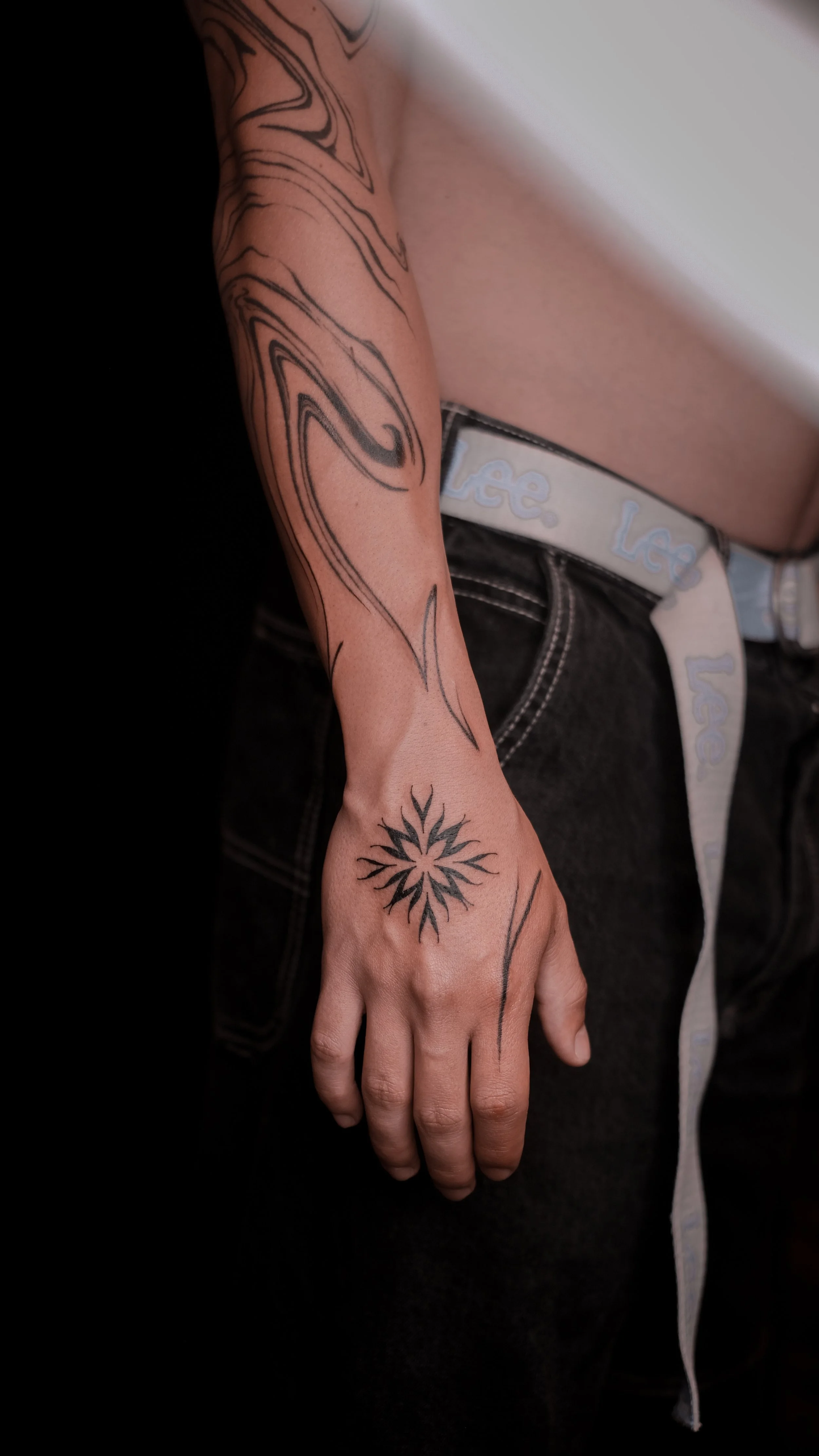 Person's arm with abstract tattoos and sun design on hand, wearing black pants and white belt with "Lee" branding.
