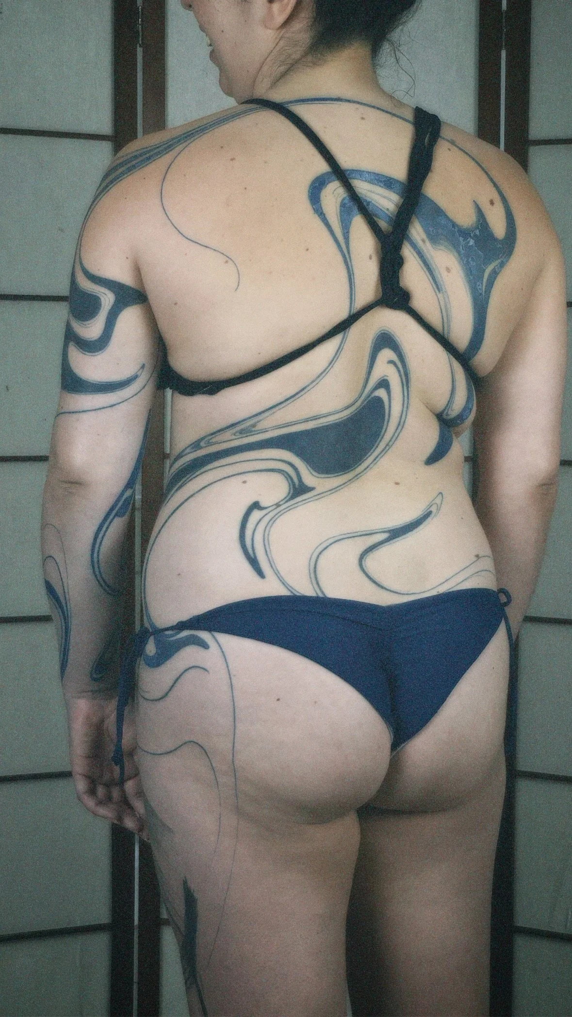 Back view of a woman with black swimwear and swirling blue and black tattoos covering her back, arm, and leg.
