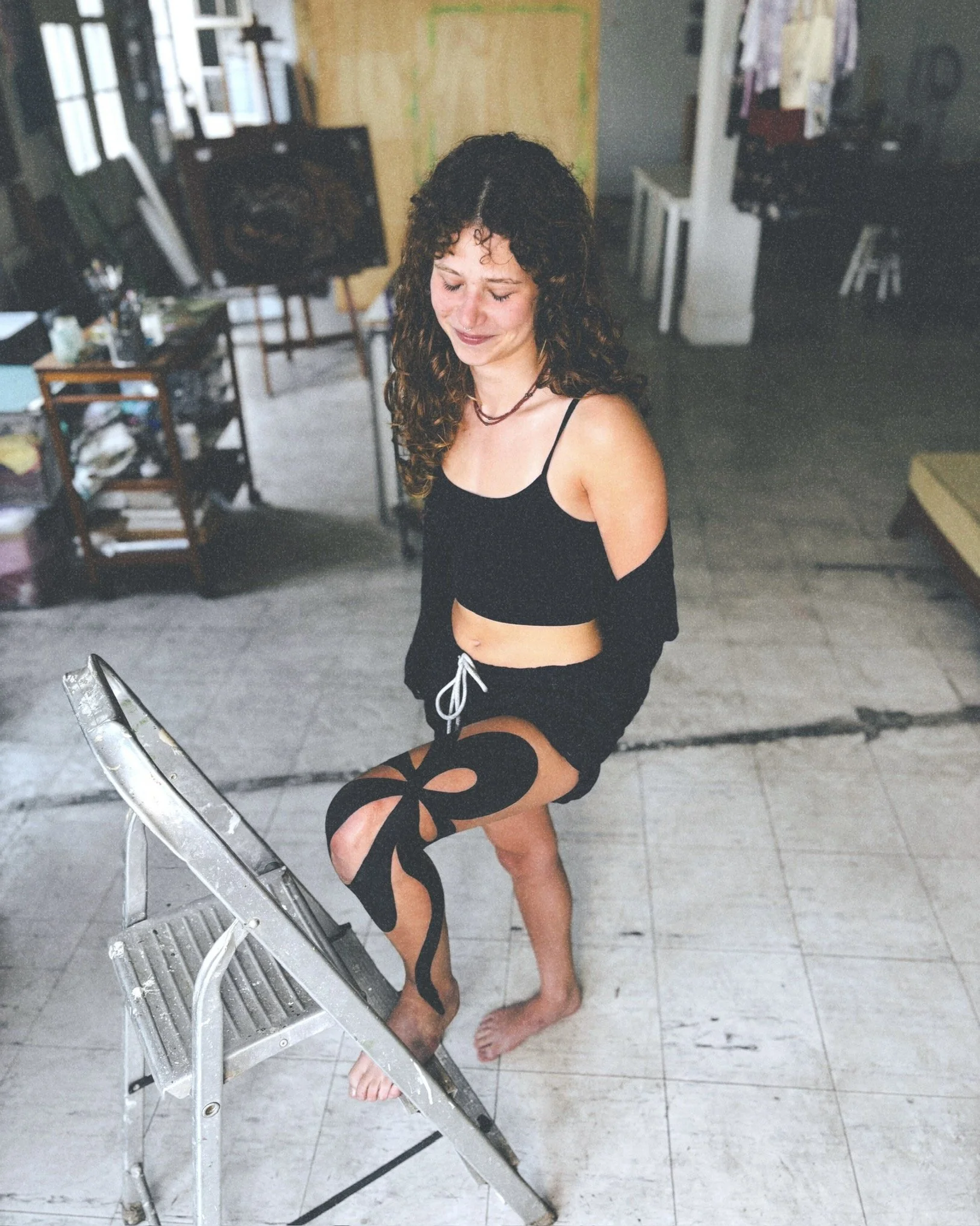 A woman with curly hair in a black crop top and shorts stands on a step stool, smiling, with one foot on her knee showing a large black ribbon tattoo on her thigh in an artist's studio.