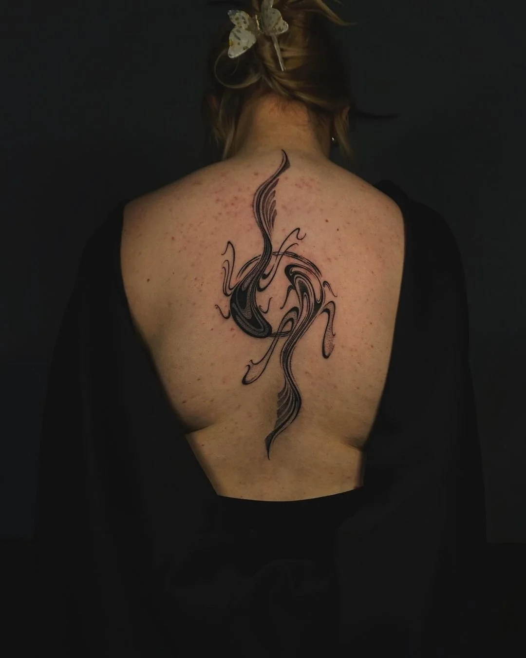 Person with abstract tattoo on their back, wearing a black garment, with hair held by a decorative clip.
