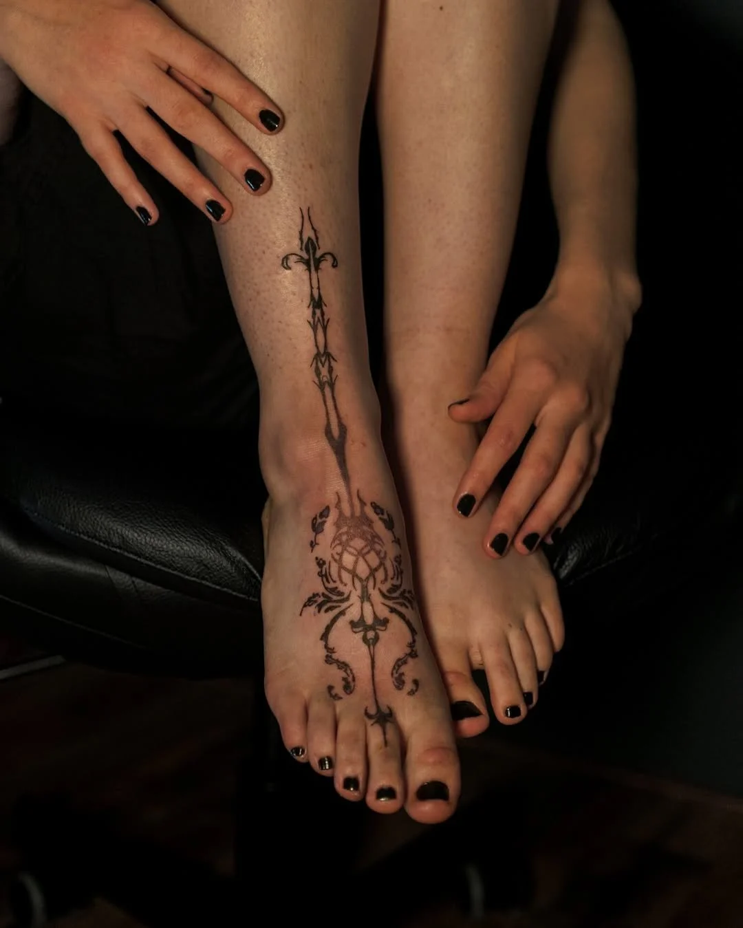 Feet with intricate black henna design and dark nail polish, accompanied by hands with matching nail color, resting on a black chair.