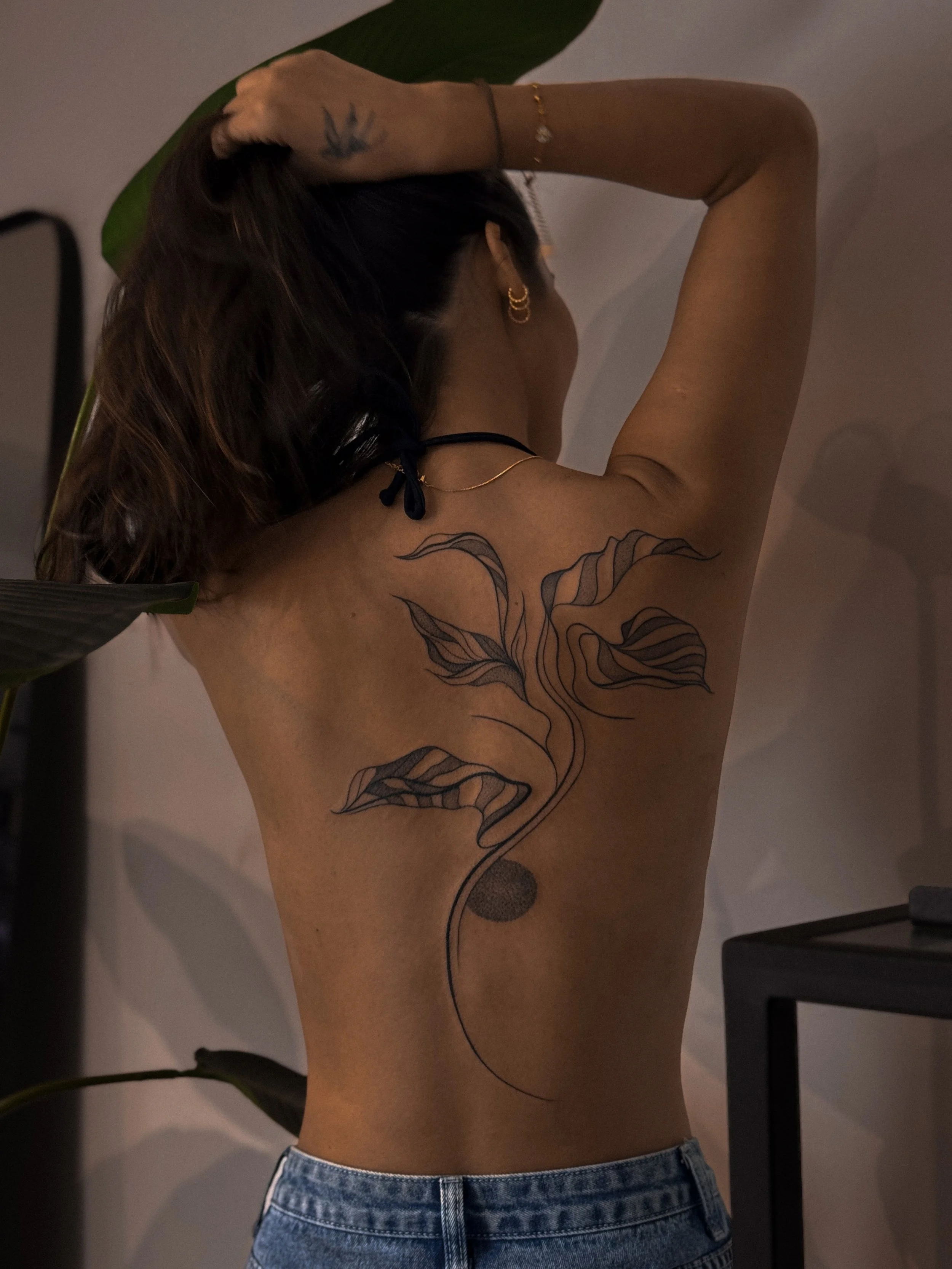 Woman with a large line art tattoo of a plant with leaves extending down her back, posing with one arm raised above her head, wearing jeans and gold jewelry, in a dimly lit room.