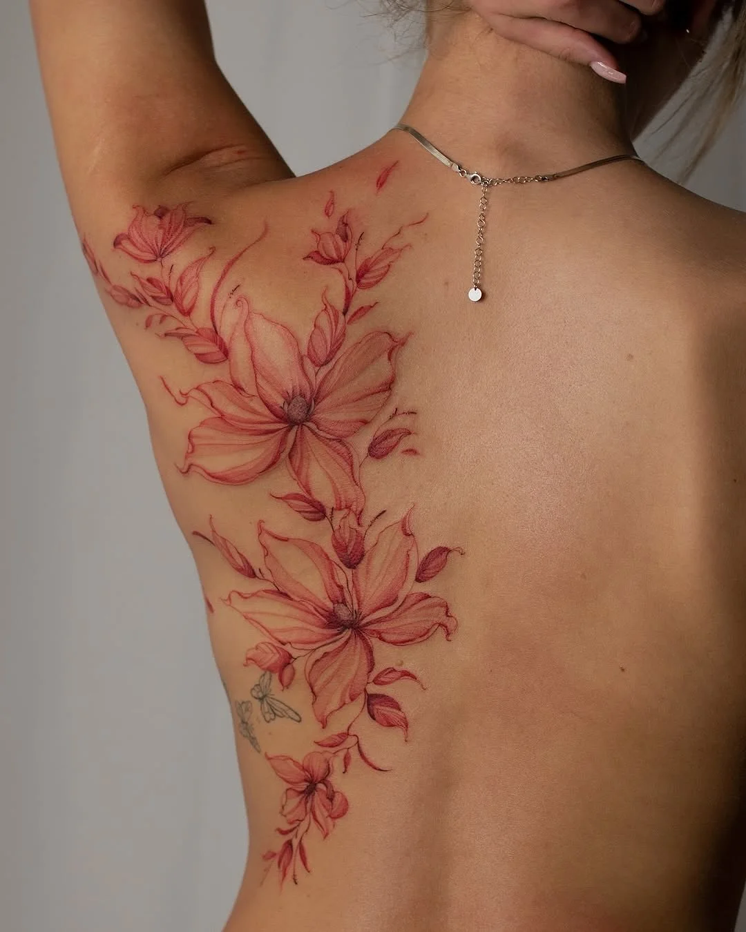 Close-up of a person's shoulder and upper chest showcasing a large, detailed tattoo of pink lilies with red accents. The person has a silver necklace with a small round pendant hanging from a chain.