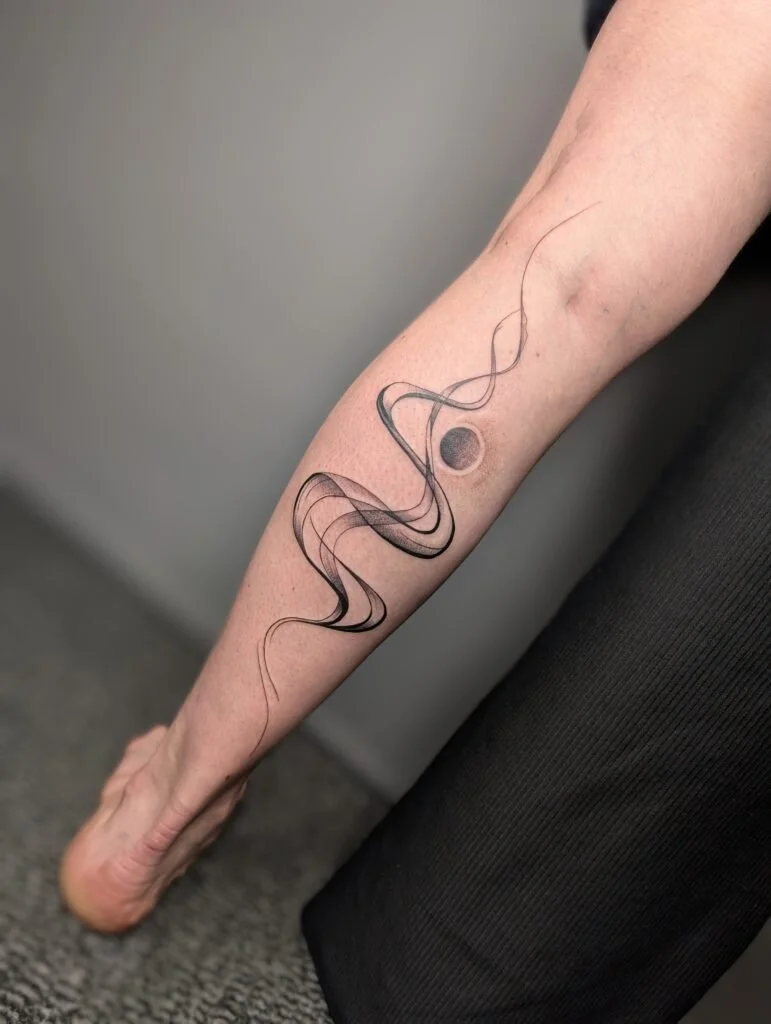 Close-up of a person's forearm with a black and gray tattoo of a swirling, flowing line design and a small solid black circle.