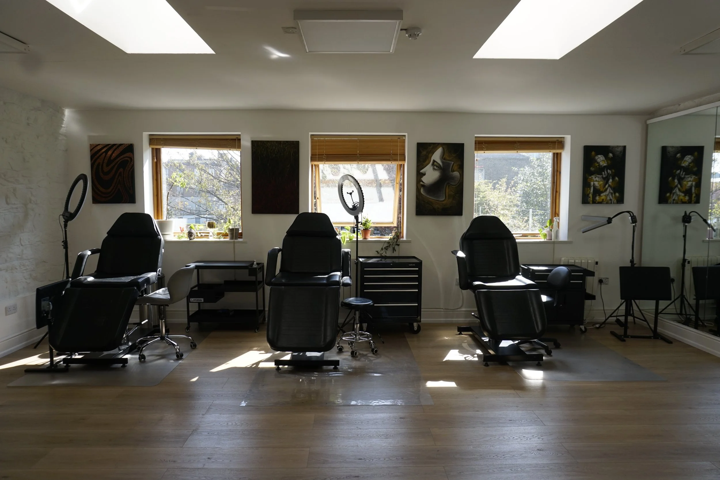 Interior of a tattoo studio with three black chairs, ring lights, artwork on walls, and a wooden floor.