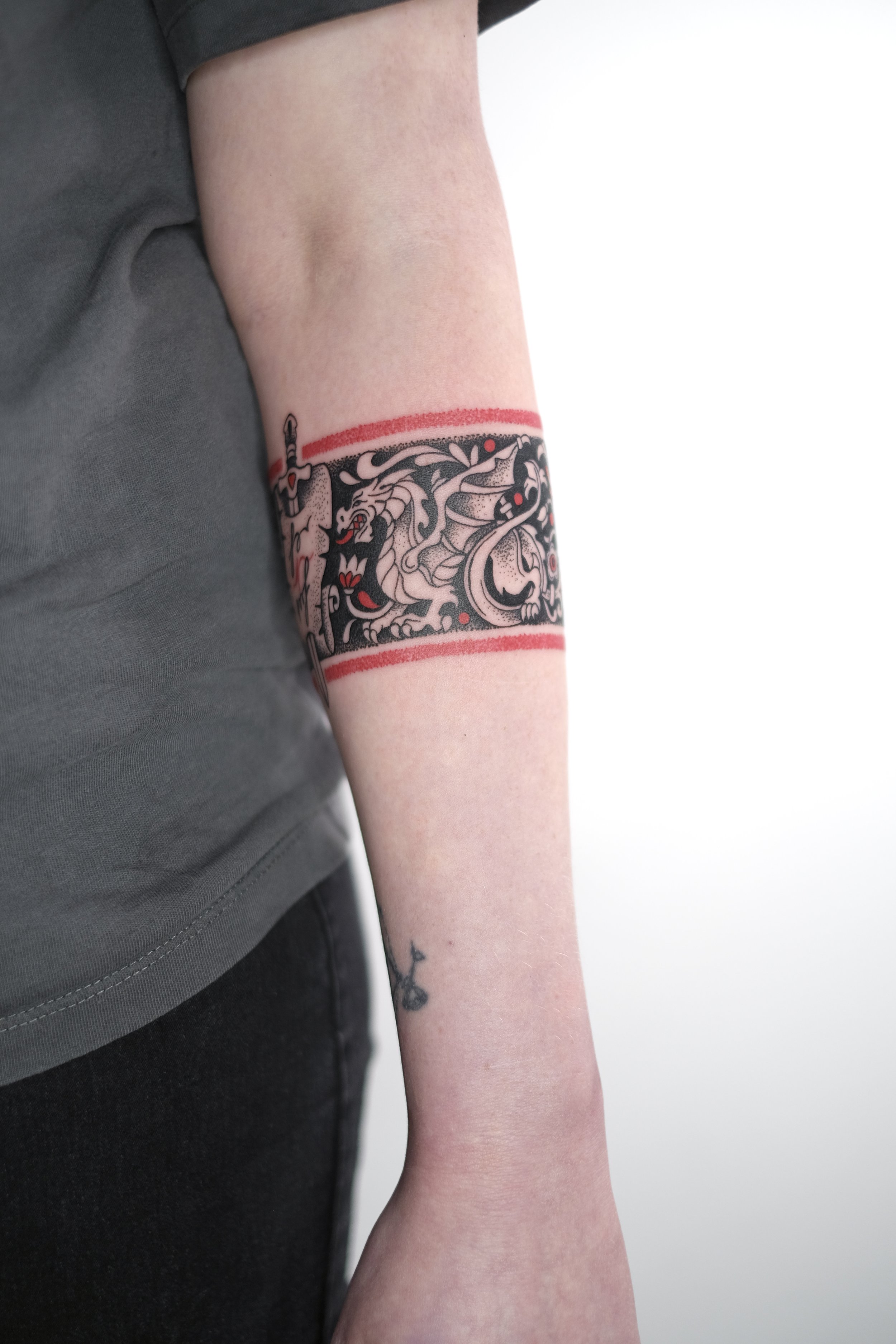 Person's arm with a large, black tattoo of a lion's face inside a rectangular border with red accents, and a small black tattoo of a sewing machine below the elbow.