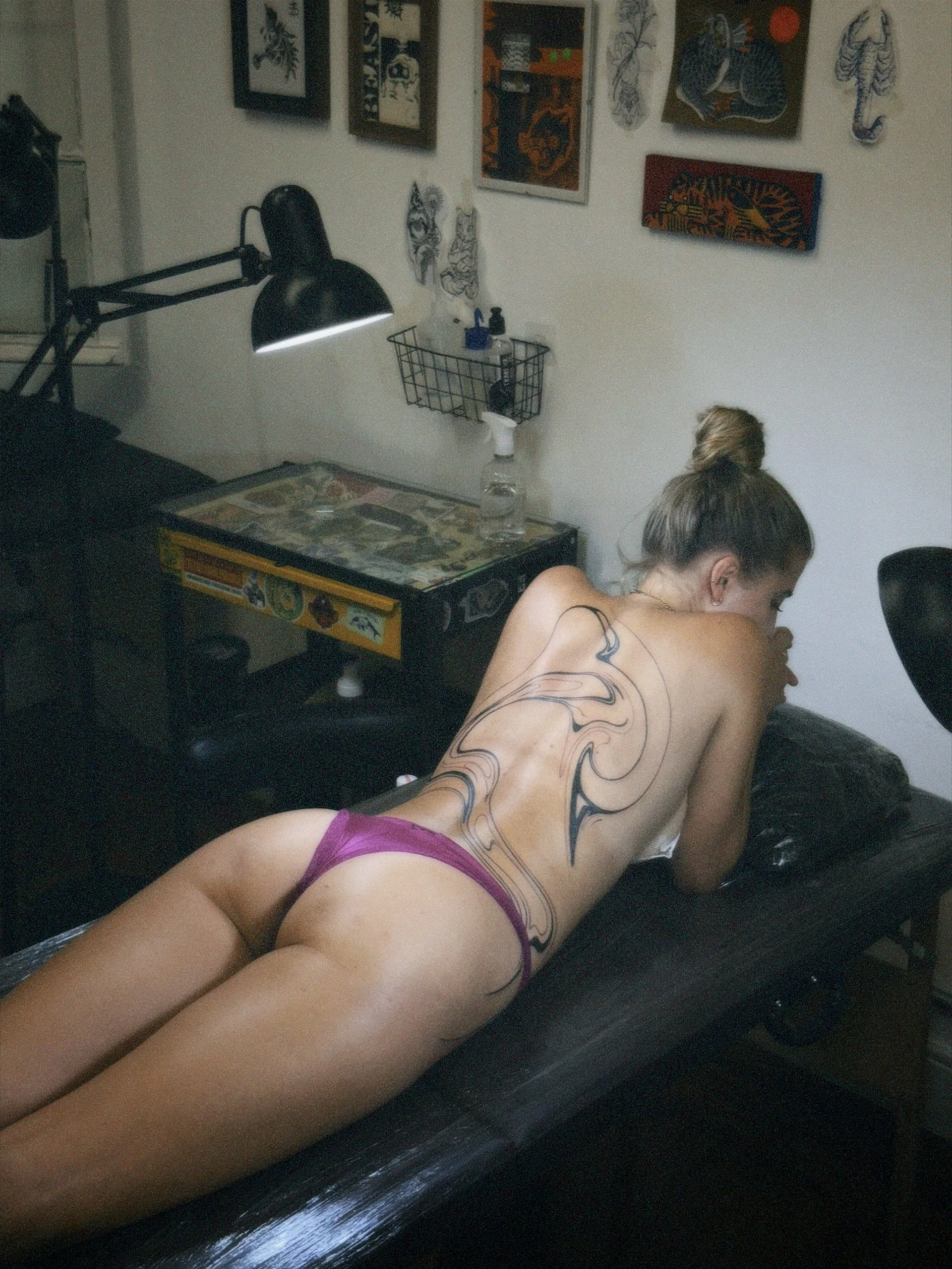 A woman with a large tattoo on her back and wearing pink underwear lies face down on a black massage table in a tattoo studio.