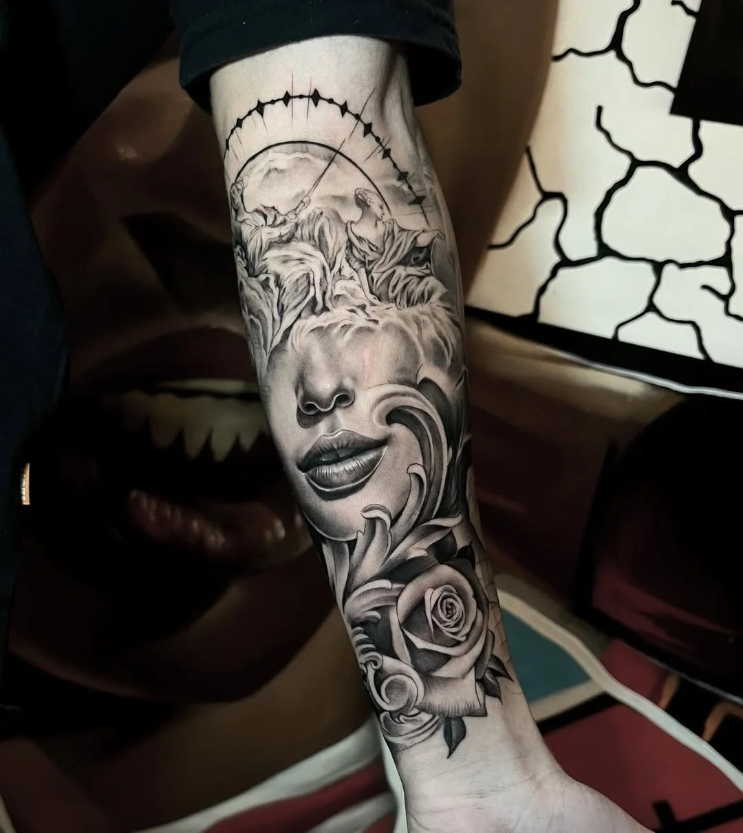 Black and gray tattoo on a person's forearm featuring a woman's face, with a rose beneath, and a landscape with mountains and clouds in the background.