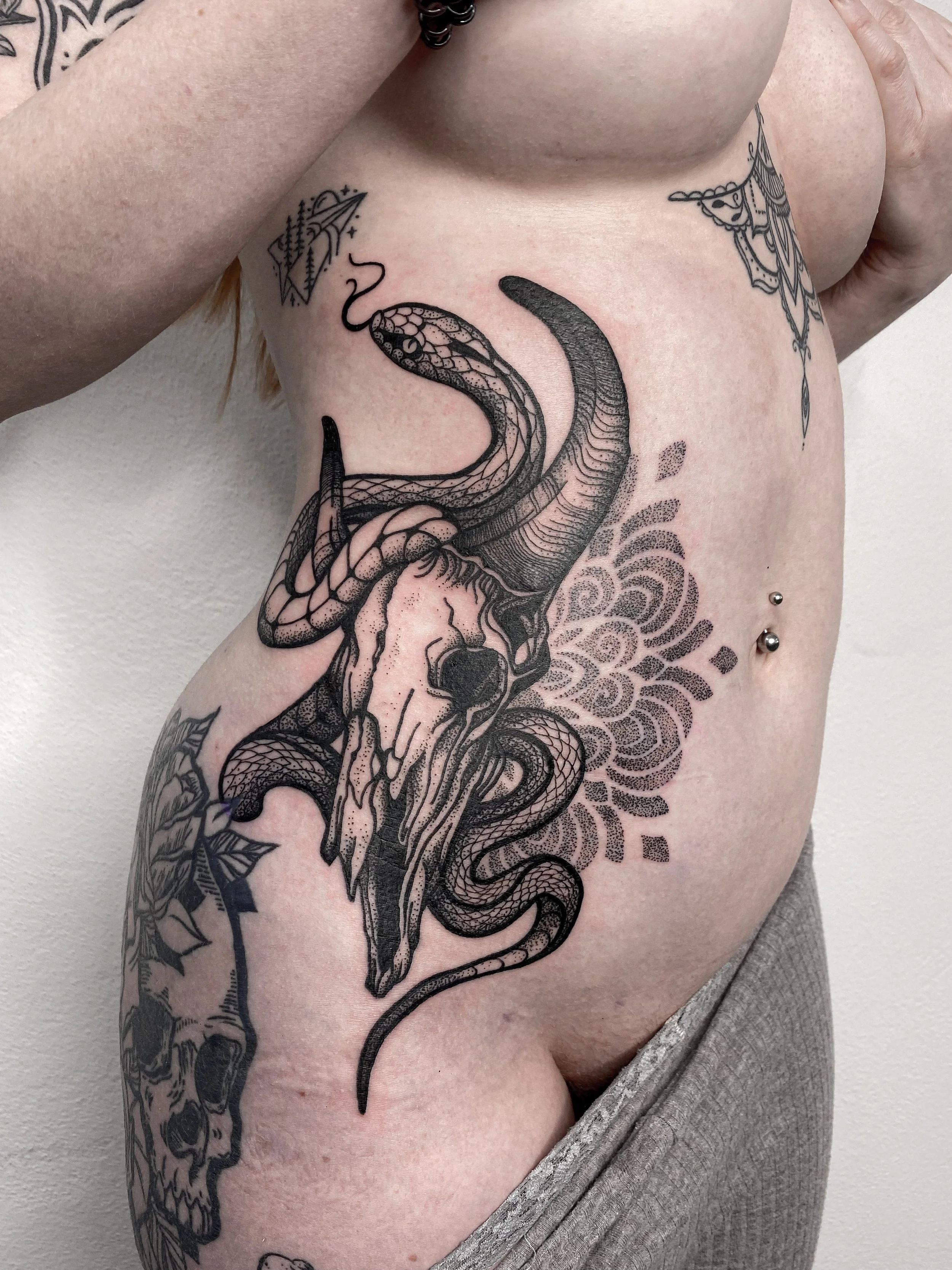 Black ink tattoo of a goat skull with twisted horns, surrounded by floral and dot patterns on person's side.