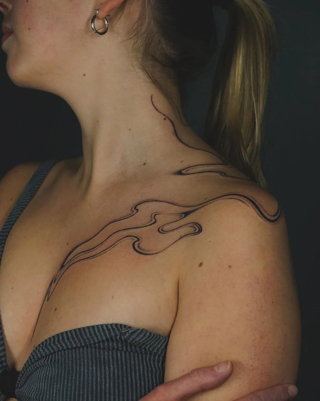 Person with abstract line tattoo on neck and shoulder, wearing a striped top and hoop earring.