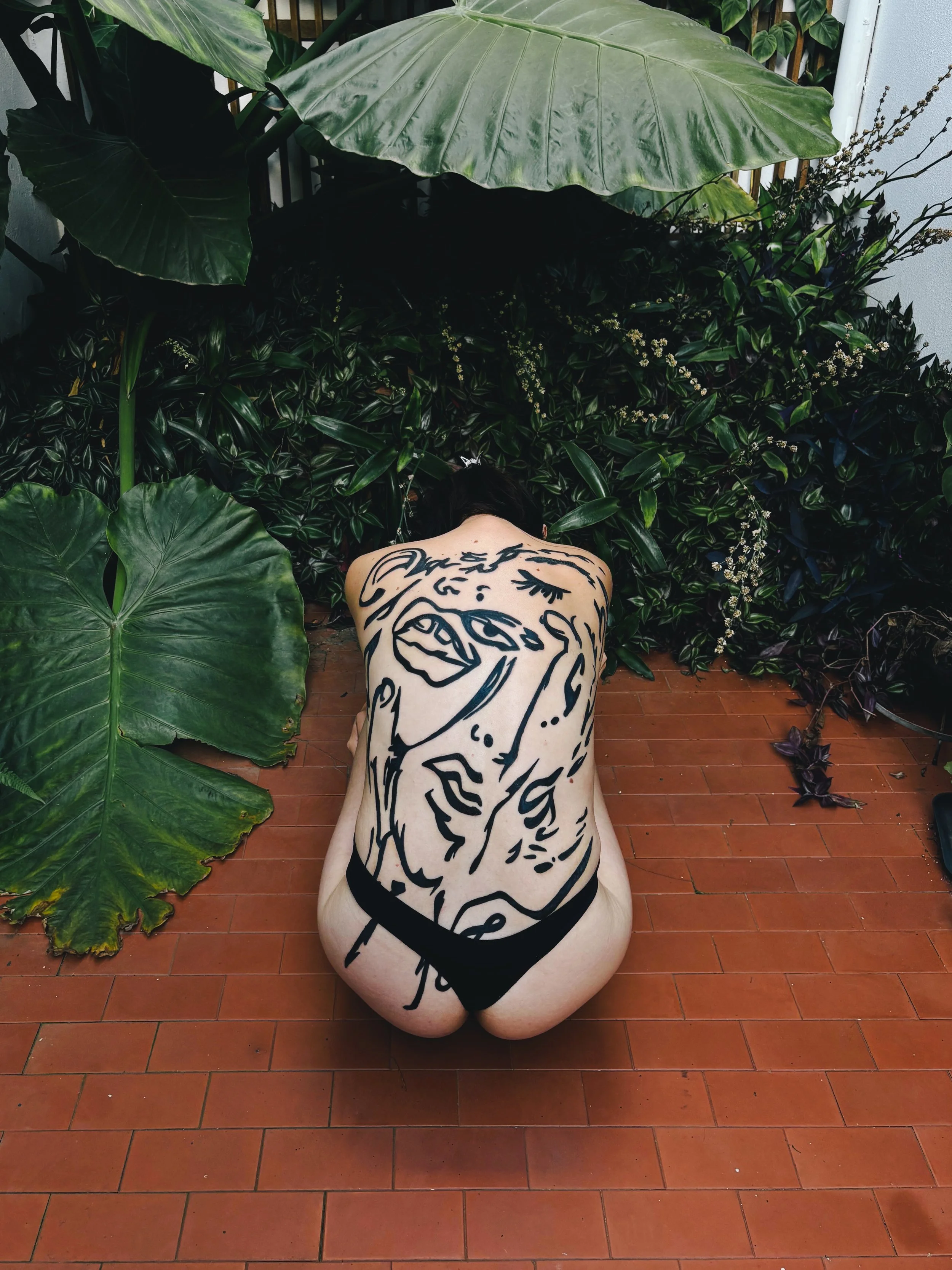 A person with a large face and abstract line art tattoo on their back, sitting on a terracotta tiled floor surrounded by lush green plants, with their head bowed.