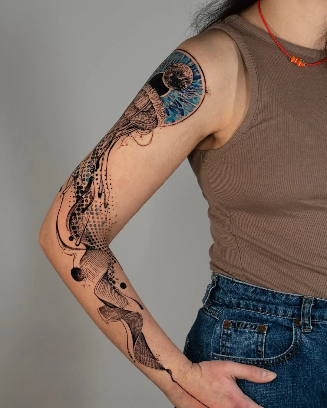 Person with detailed artistic tattoo on arm, featuring an intricate design with a large eye, ocean elements, and abstract patterns in black and blue ink. Wearing a taupe sleeveless top and blue jeans.