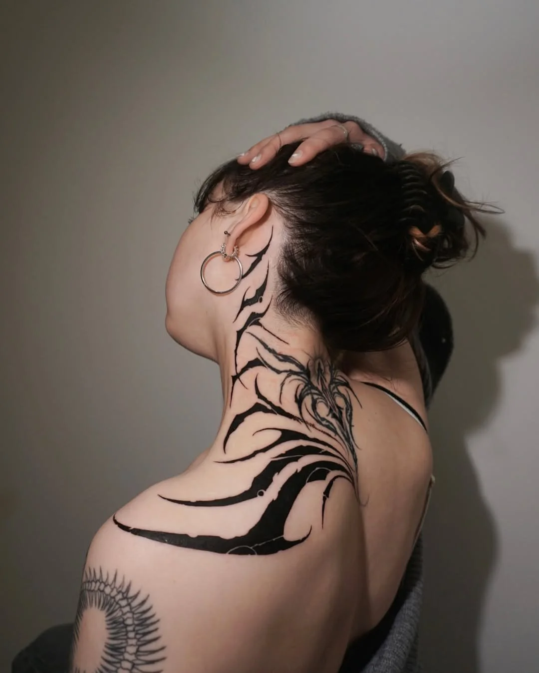 A woman with dark hair pulled up, wearing large hoop earrings, displaying a black tribal-style tattoo that covers her neck, shoulder, and part of her back, with a plain gray background.