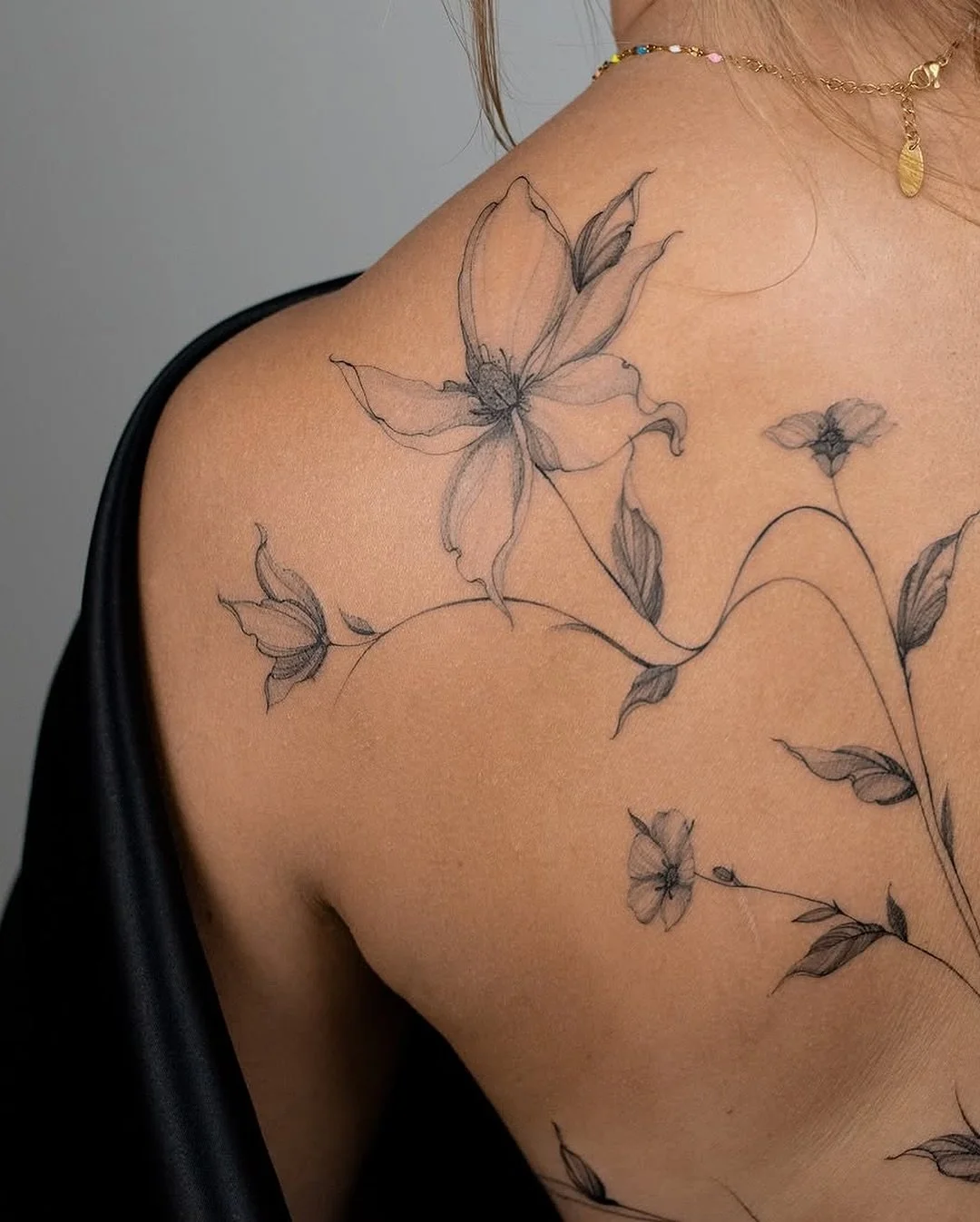 A person's shoulder and upper arm with a floral tattoo of delicate, black-outlined flowers and leaves.