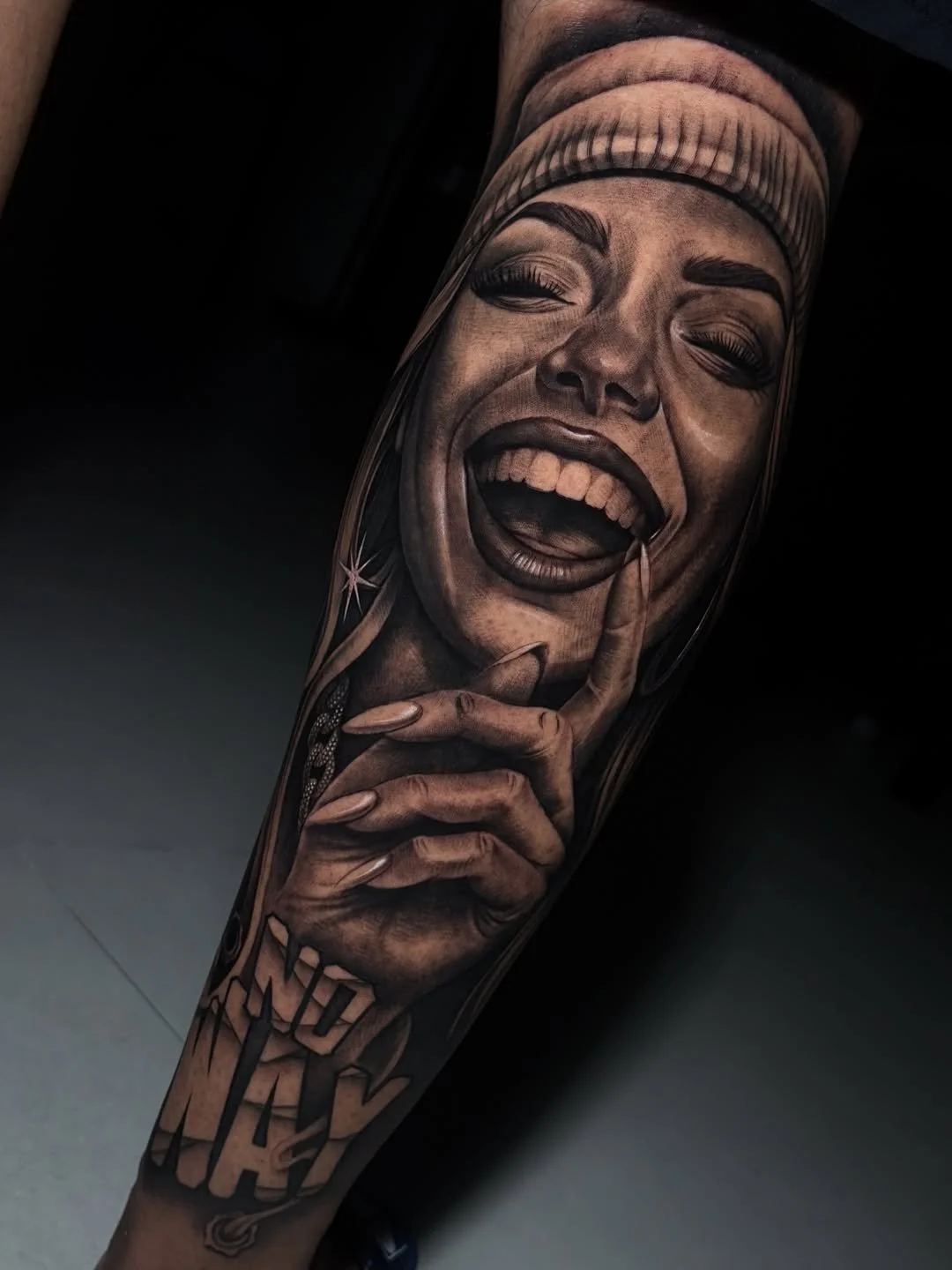 A detailed black and gray realism tattoo of a woman laughing, with her eyes closed and finger on her lips, on a person's arm.