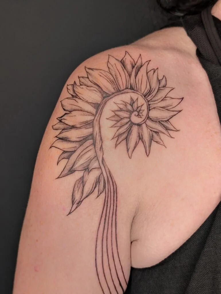 Black and gray tattoo of a sunflower with a spiral shell in the center on a person's shoulder.