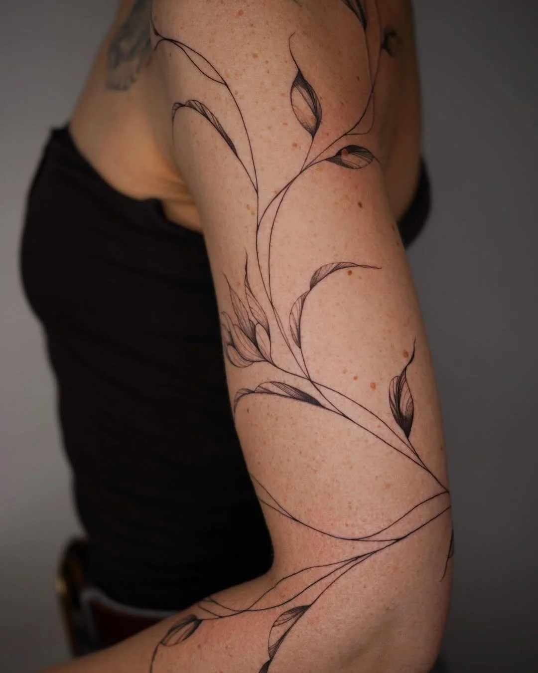 A person's arm with a black ink tattoo of a delicate floral design featuring thin, flowing stems and leaves.