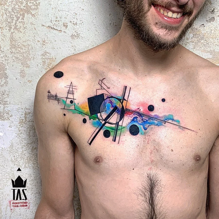 A man with a colorful geometric tattoo on his chest, featuring abstract shapes and lines on a textured wall background.