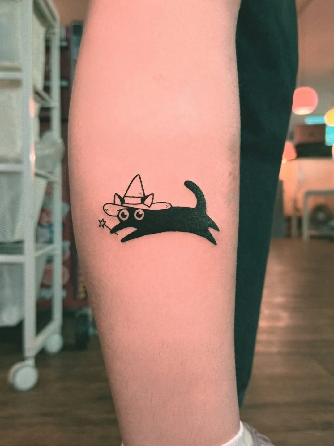 Tattoo of a black cat wearing a witch hat, with wide eyes and a small star wand, on a person's forearm.
