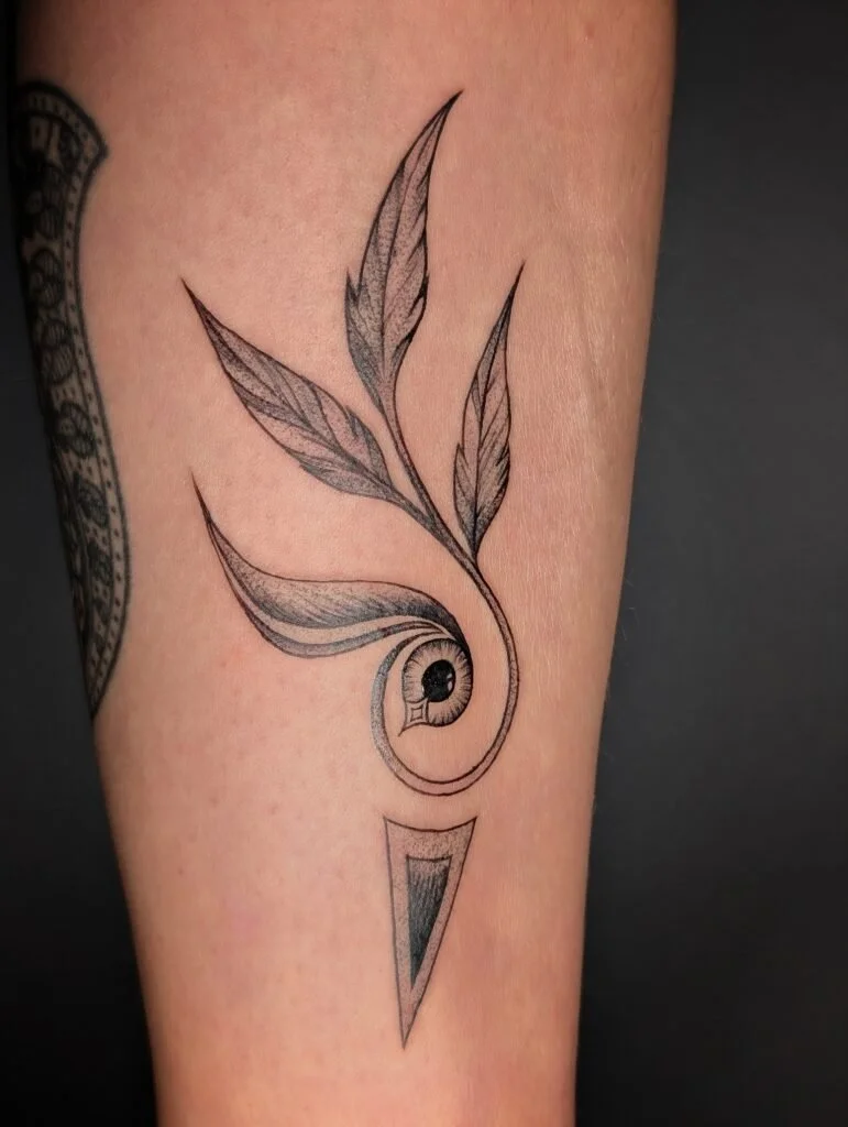 A black and gray tattoo of a stylized eye within a curl, with four elongated, detailed leaves extending from it, and a triangular shape below.