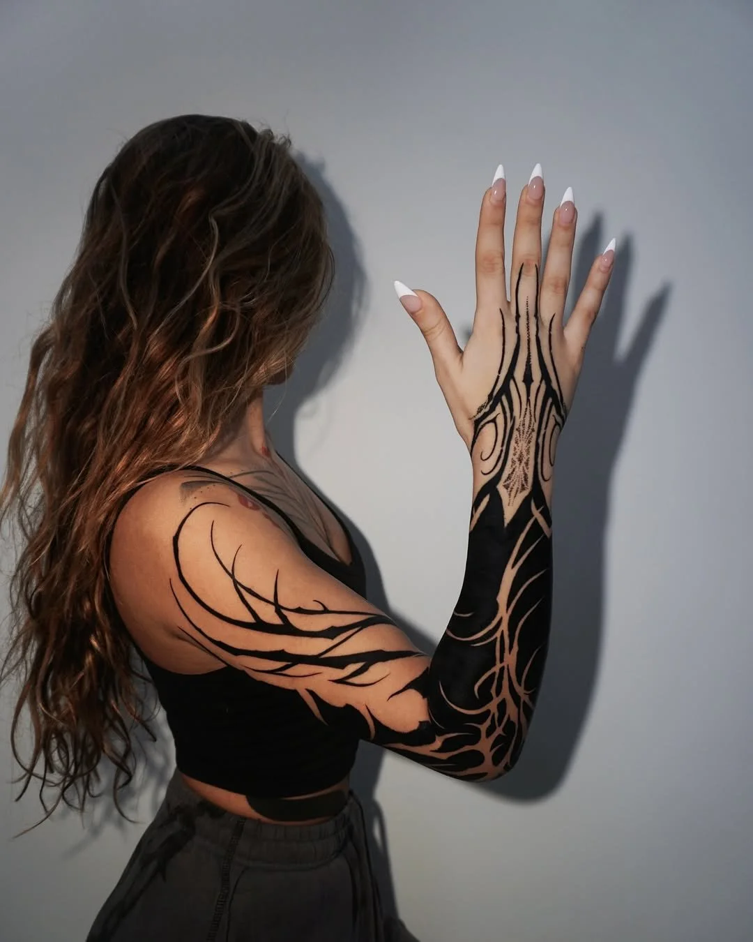 A woman with long, wavy brown hair standing against a plain wall, showing her tattoos on her arm and hand. She is wearing a black sleeveless top and dark pants, with her hand raised and fingers spread.