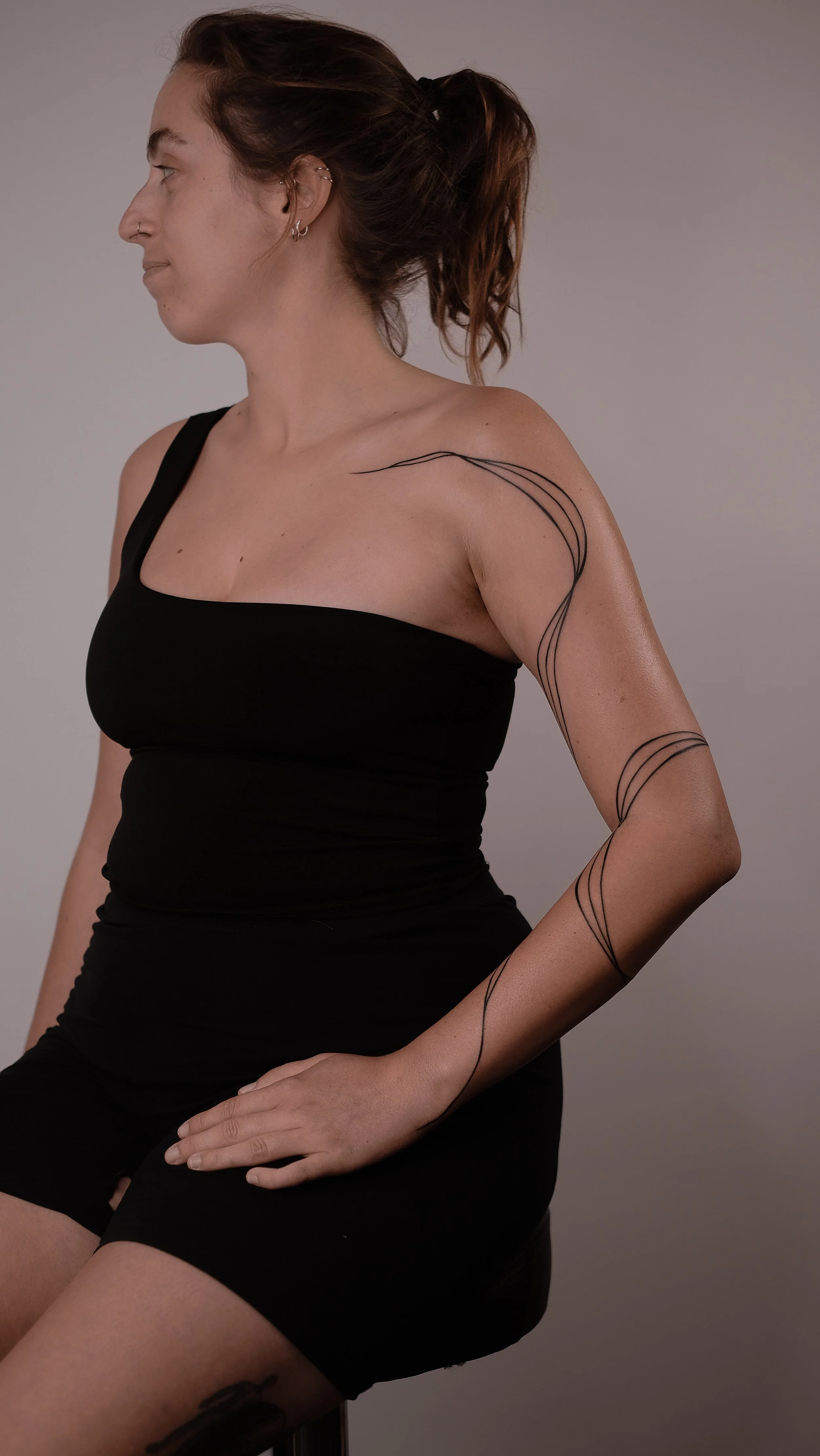 Woman with a ponytail wearing a black one-shoulder top, displaying abstract line tattoos on her right arm and shoulder, seated on a stool against a gray background.