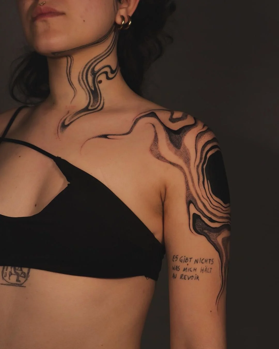 Person wearing a black top with intricate black abstract tattoos on the neck and shoulder. The tattoos feature flowing, wavy lines. There's additional text tattooed on the upper arm in a different style.