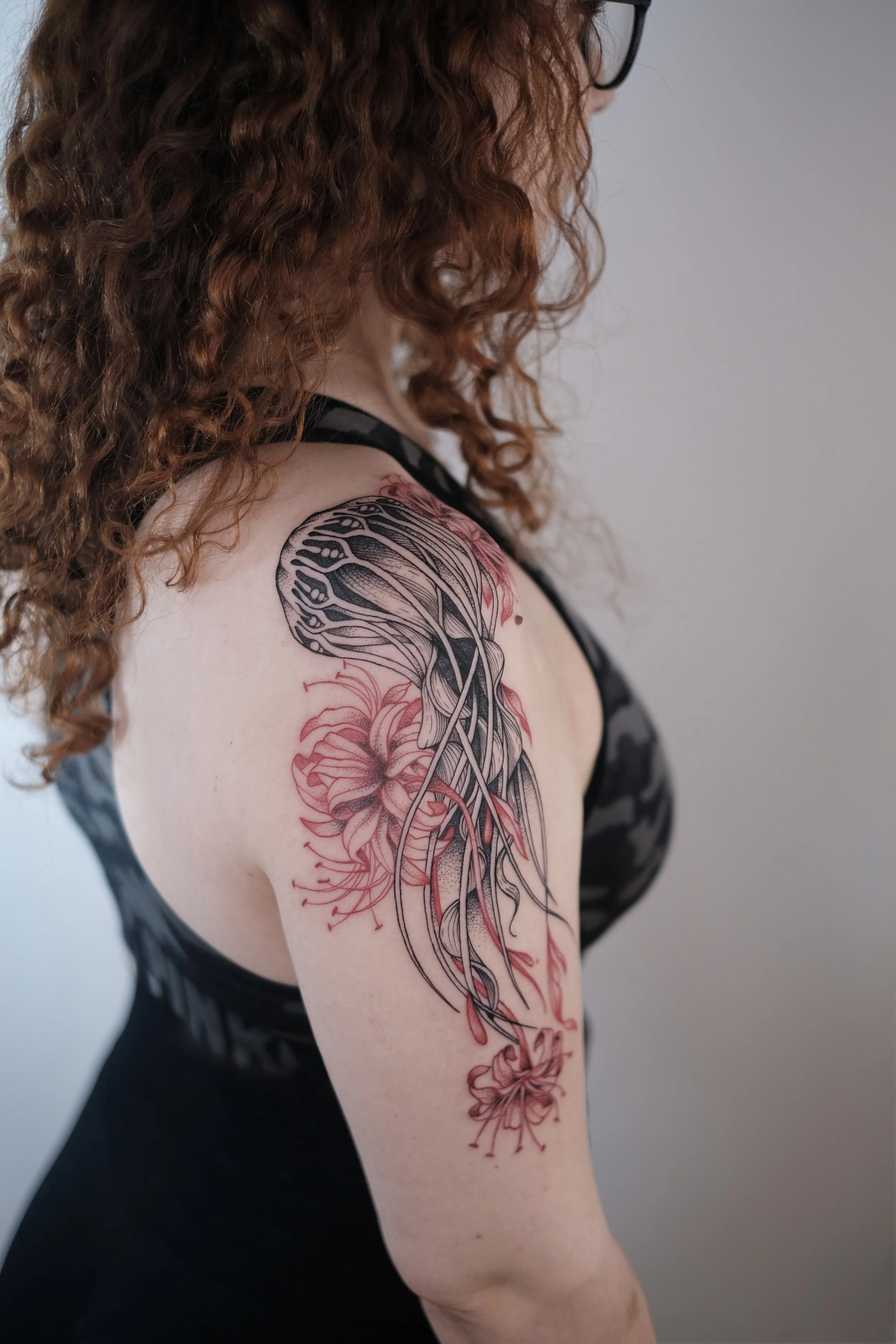 A woman with curly red hair and glasses, wearing a black tank top, has a large tattoo on her upper arm featuring black and red floral and abstract designs.