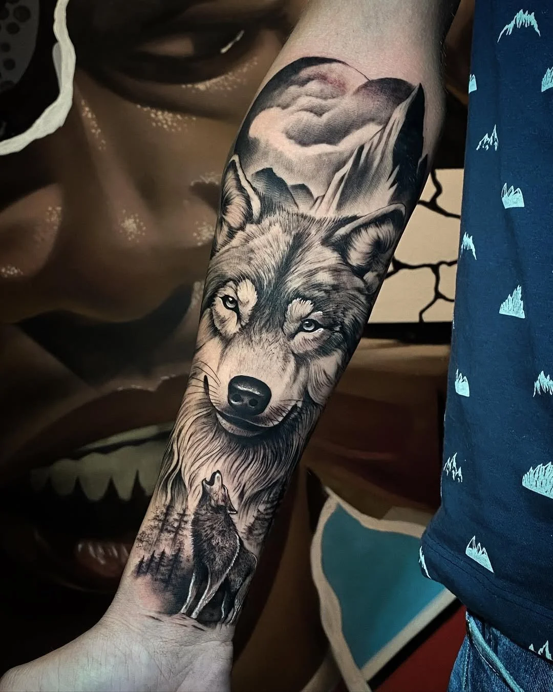 Black and gray realistic tattoo of a wolf and a howling wolf in a forest with mountains and clouds in the background on a person's arm.
