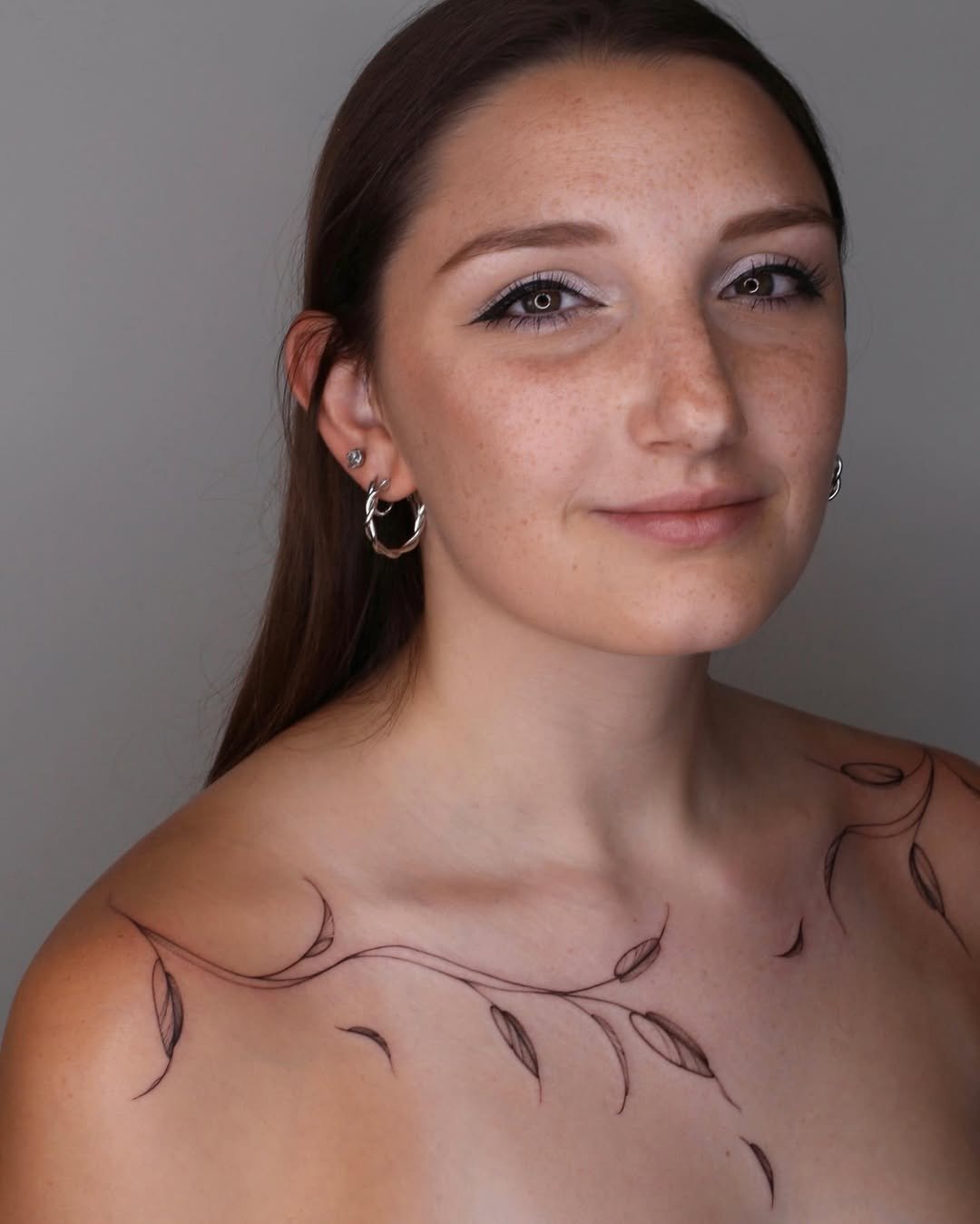 A young woman with brown hair and freckles, wearing earrings, has tattoos of a vine with leaves on her shoulder.