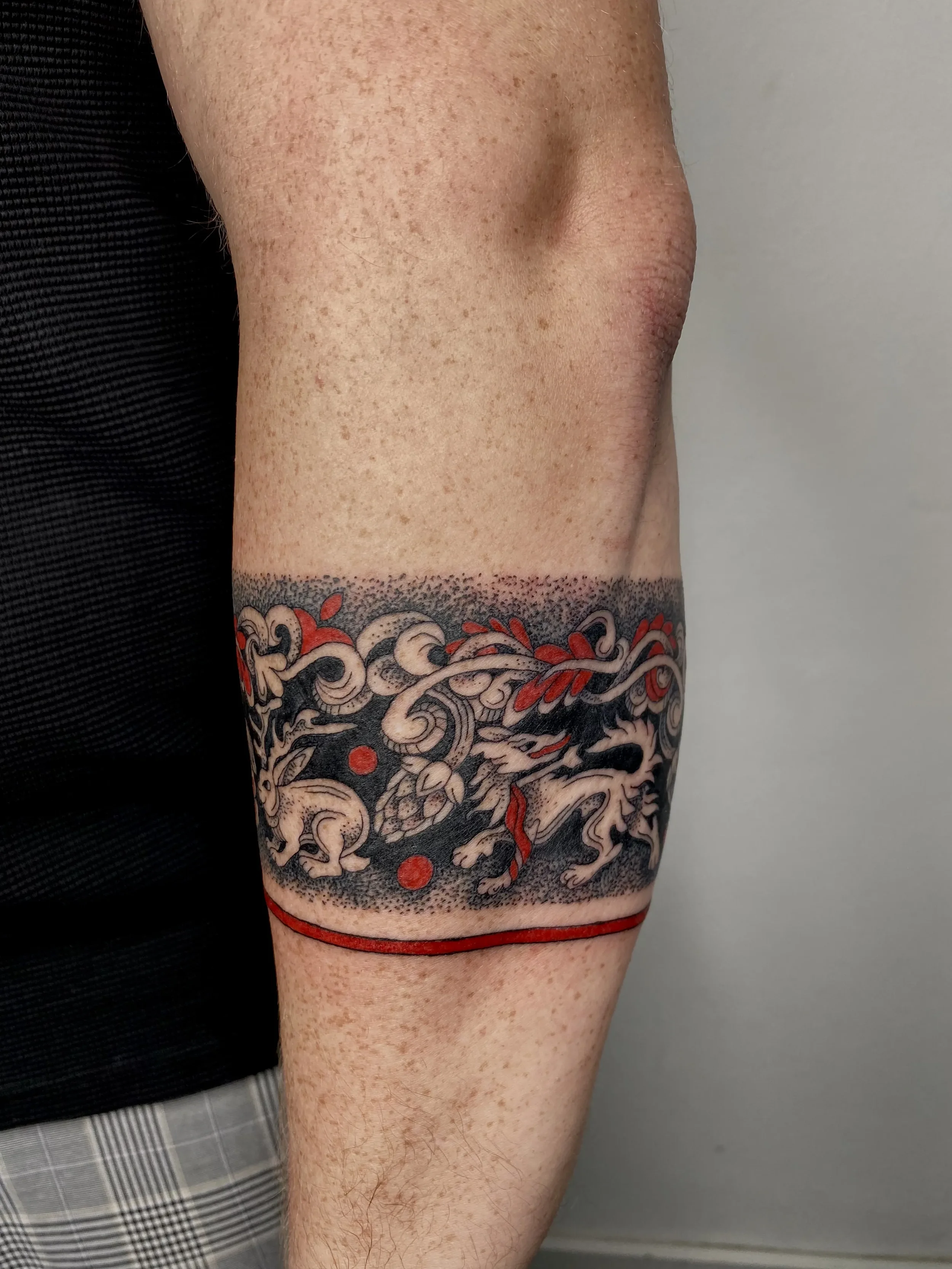 Tattoo of a lion surrounded by decorative elements, with a horizontal red line and a red upper border on a person's upper arm.