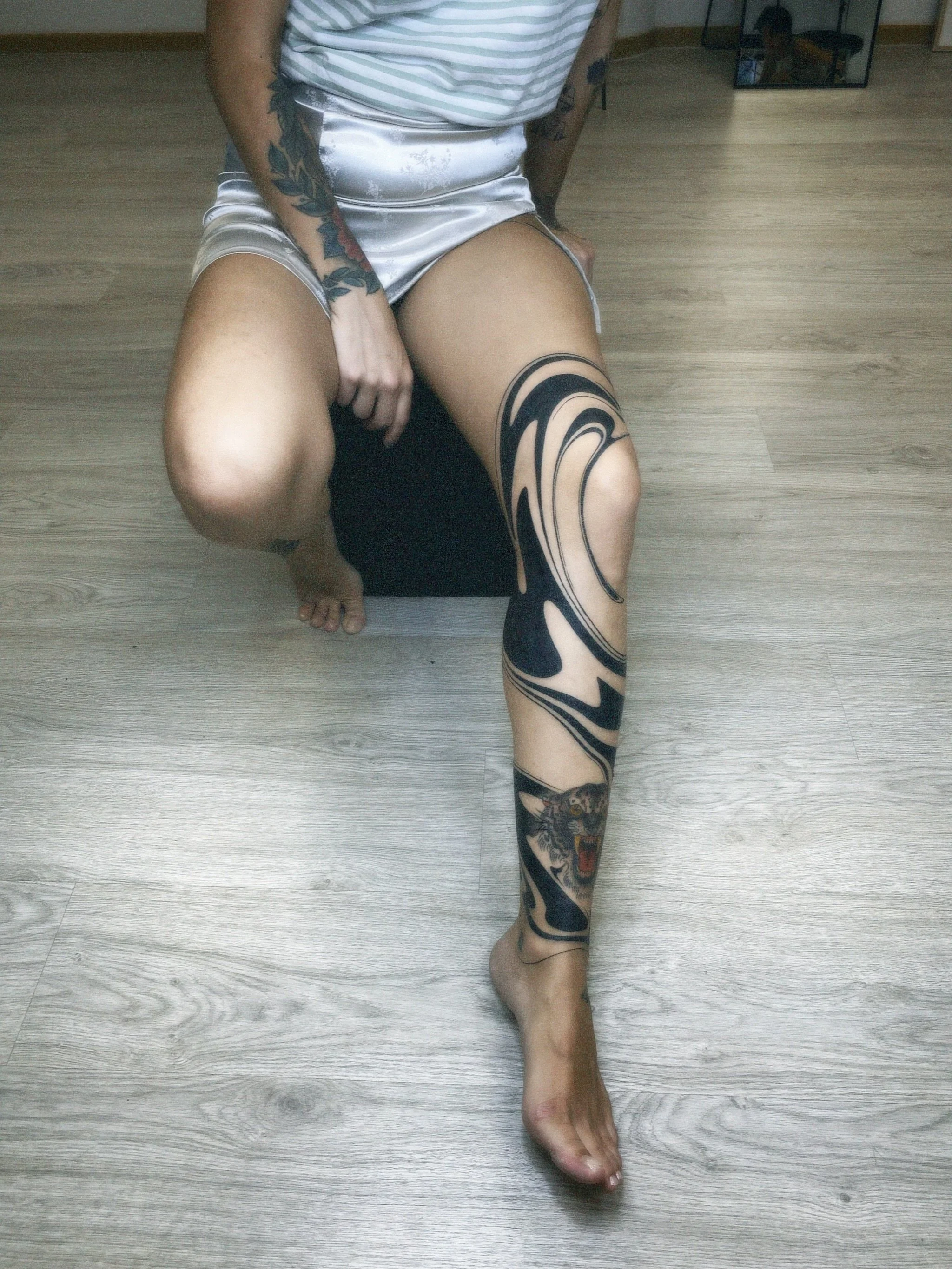 Person sitting on a black stool with tattoos, wearing satin shorts and a striped shirt, with a large black and gray tribal and tiger tattoo on their right leg, on a wood floor.