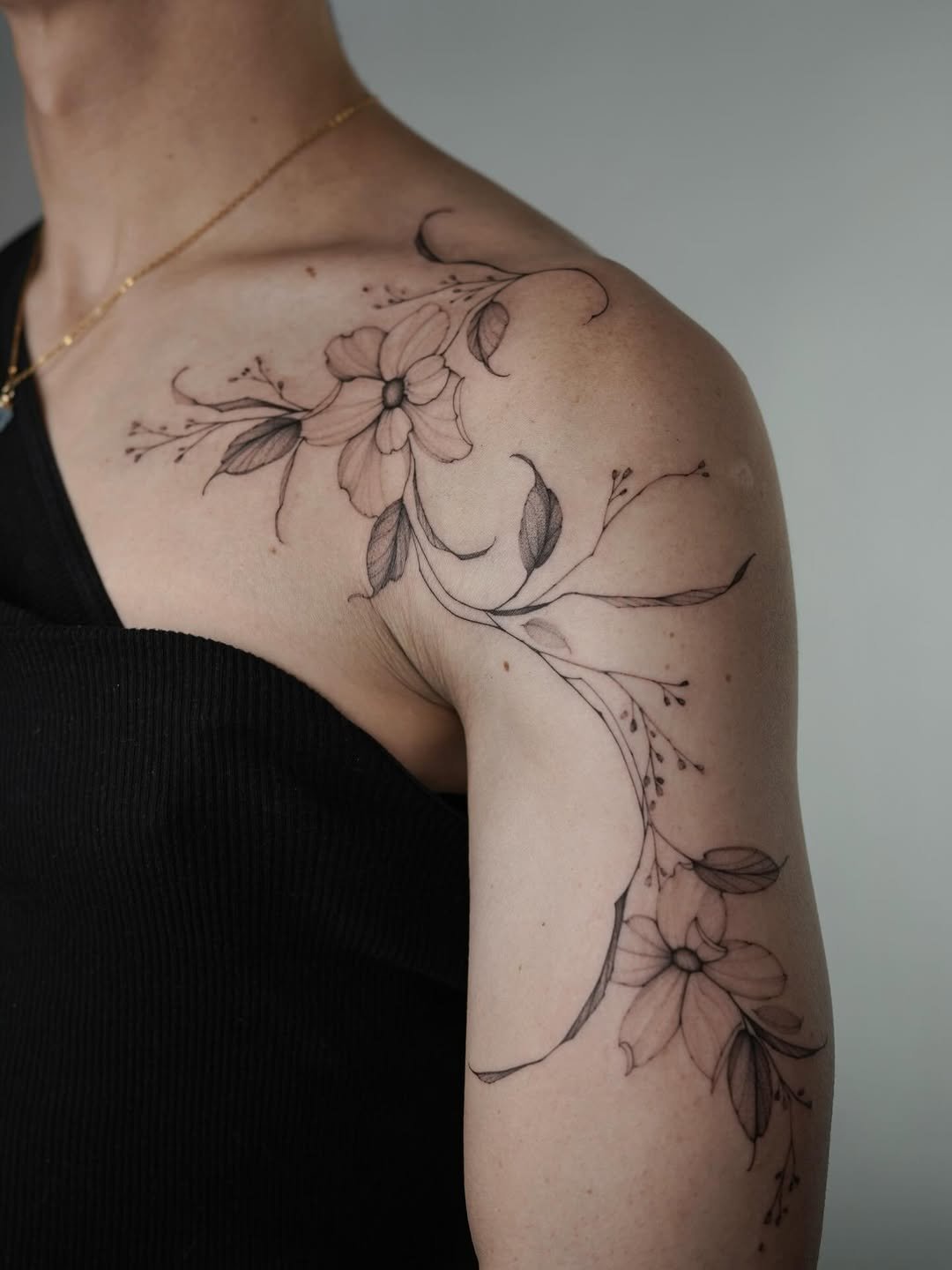 A black floral tattoo on a person's shoulder and upper arm featuring outlined flowers, leaves, and vines.