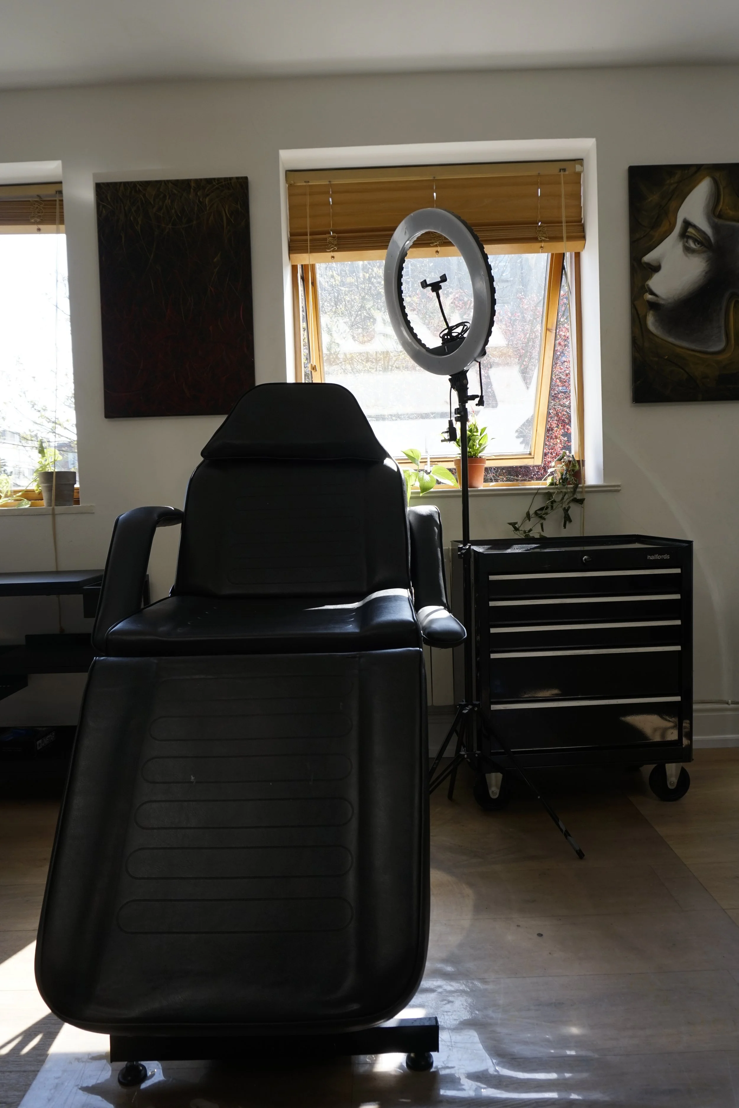 Tattoo studio interior with a black chair, ring light, wooden blinds, tool chest, and artwork on walls.