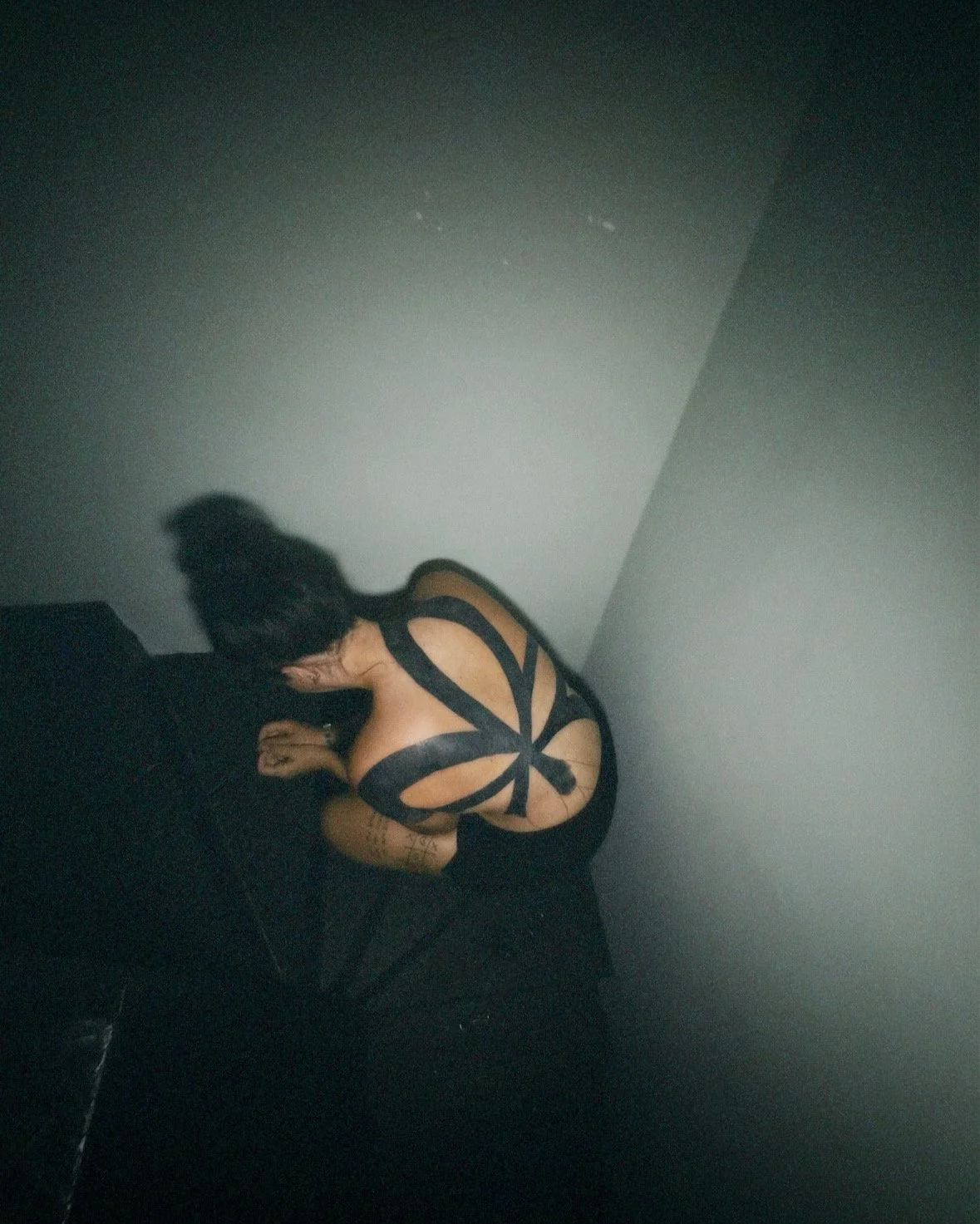 A woman in black lingerie with a butterfly-shaped back design, sitting on a dark bed against a plain wall.