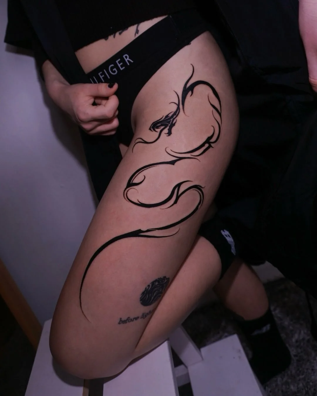 A person's thigh with a large black tribal dragon tattoo, partially covered by black underwear and black shorts, with the word 'Tommy' visible on the waistband.