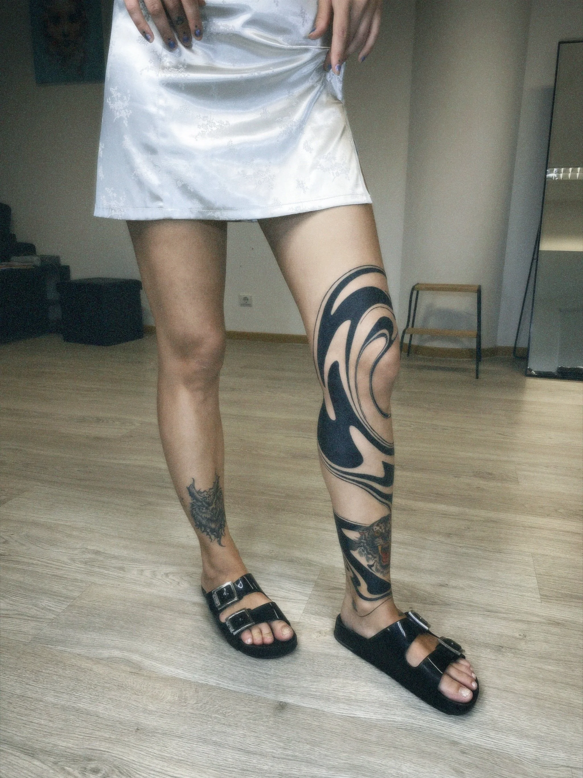 Person with tattoos on legs wearing black slides and a shiny silver skirt, standing on wooden floor, partly visible in a room.