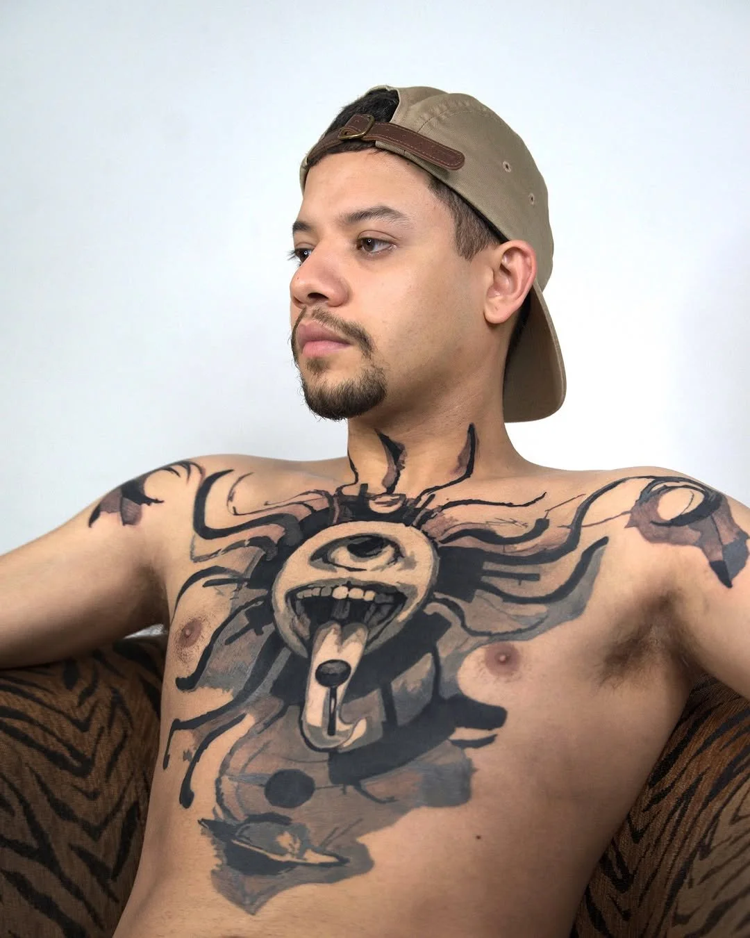 Man with a large abstract tattoo featuring an eye and tongue on his chest, wearing a backward baseball cap.