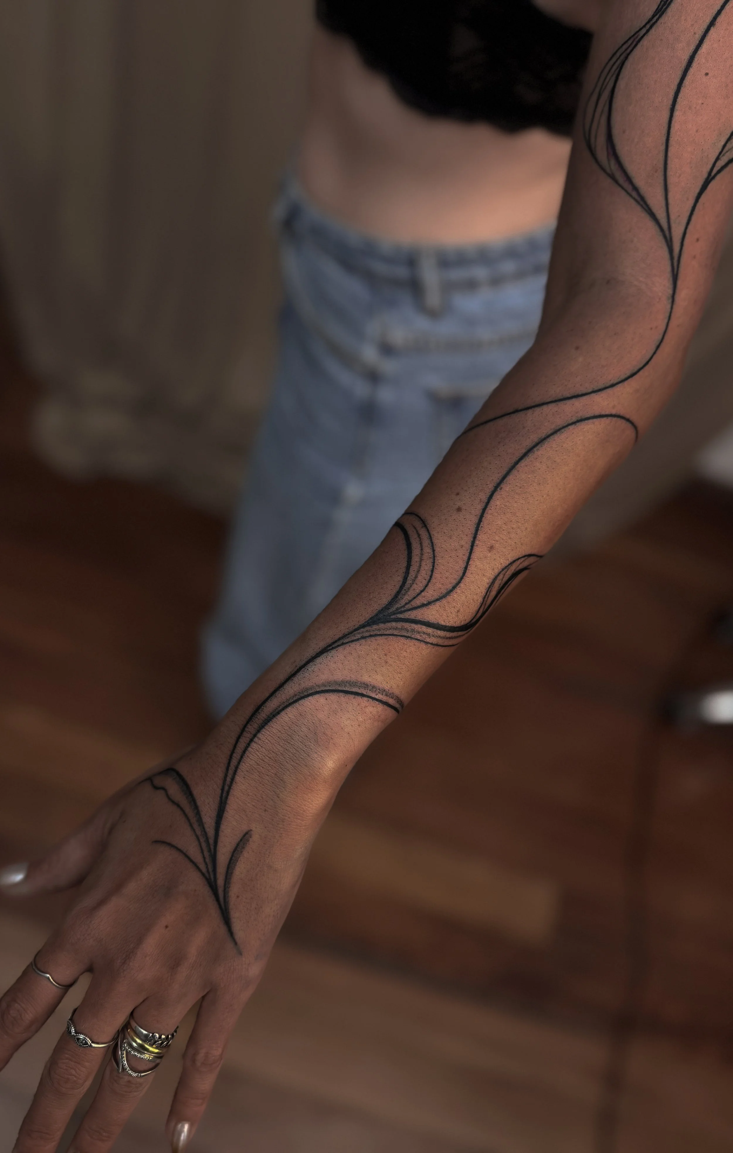 A person's arm with a black abstract line tattoo, partially visible, with a background of a wooden floor and a blurred out person wearing jeans and a top.