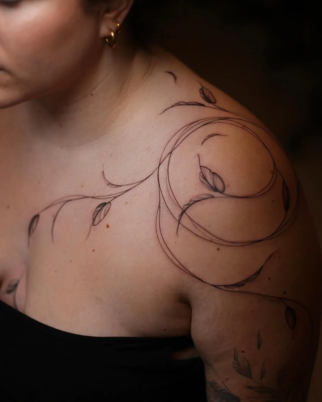 A person's shoulder and upper chest with a floral tattoo outline and some earrings.