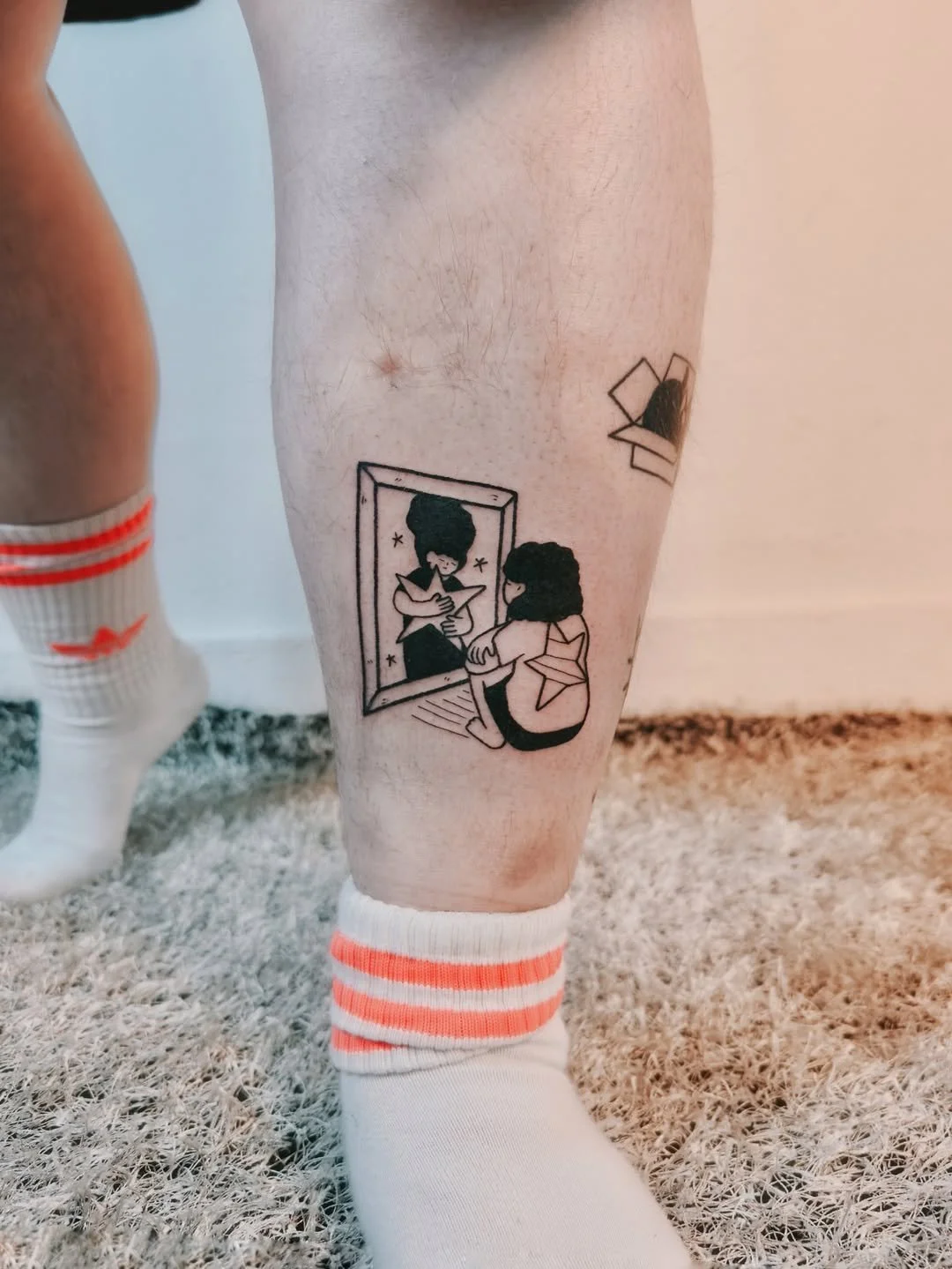 Black tattoo of a girl sitting by a mirror looking at her reflection, with a star on her shirt, and another tattoo of a person with a star on their back on the person's thigh.