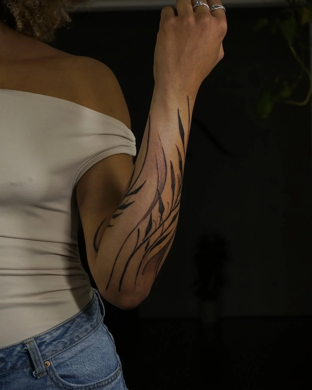 Person with abstract black line tattoo on forearm, wearing white off-shoulder top and denim jeans, against dark background.