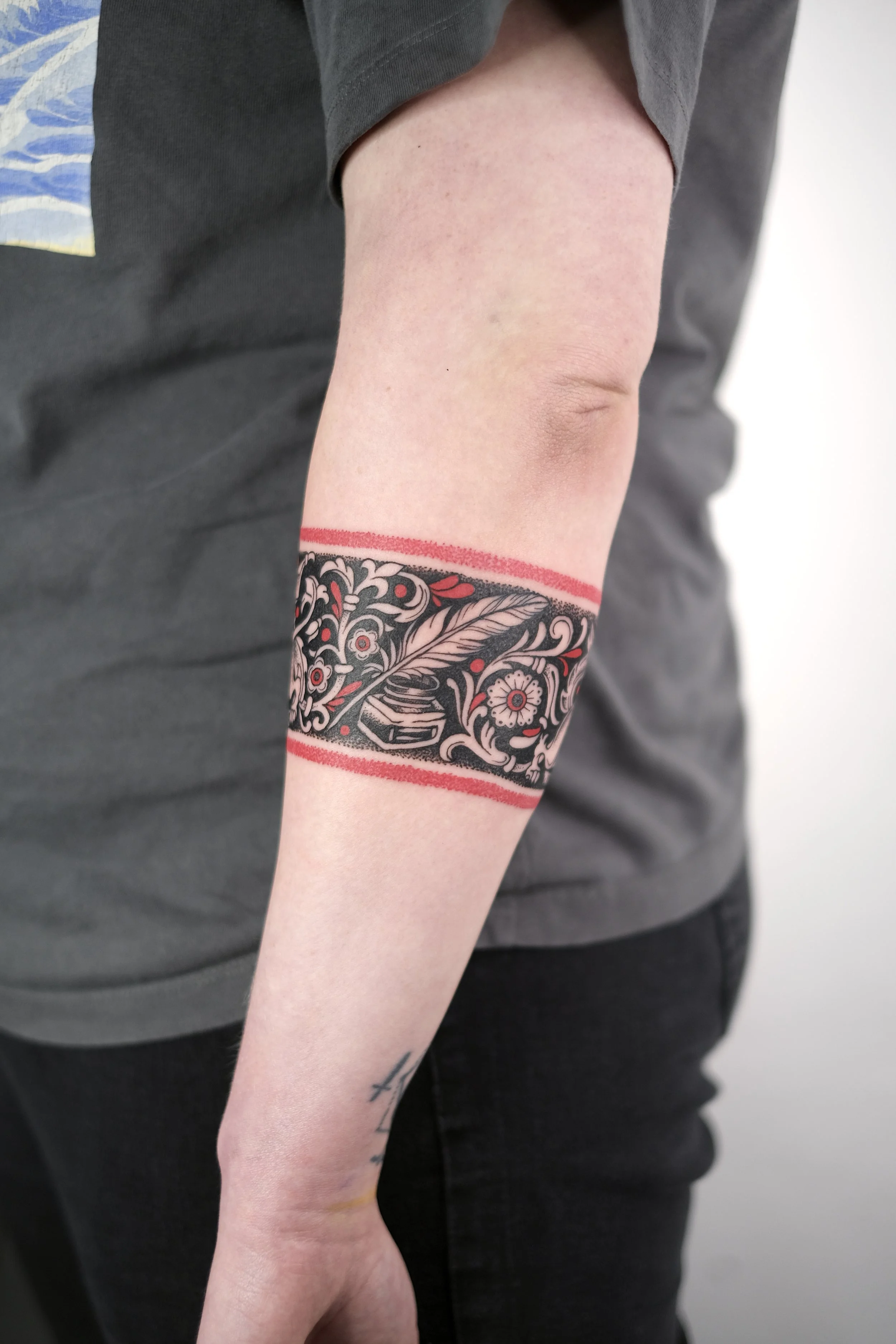 Close-up of a person's forearm with a detailed black and red floral tattoo band, wearing a gray shirt and black pants.