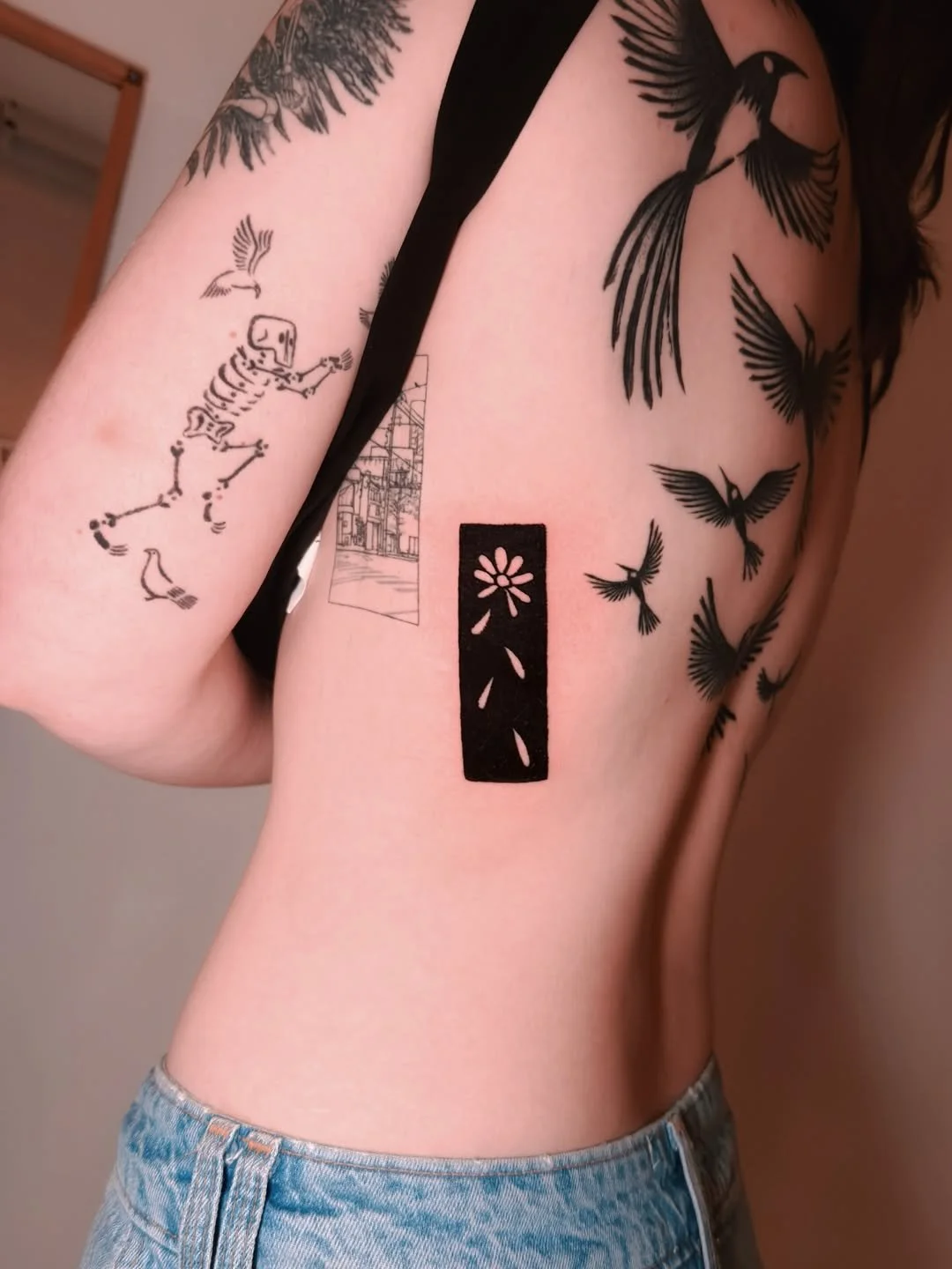 Tattoos on person's arm and side, including a skeleton dancing, a woman sitting on a chair, and flying birds.