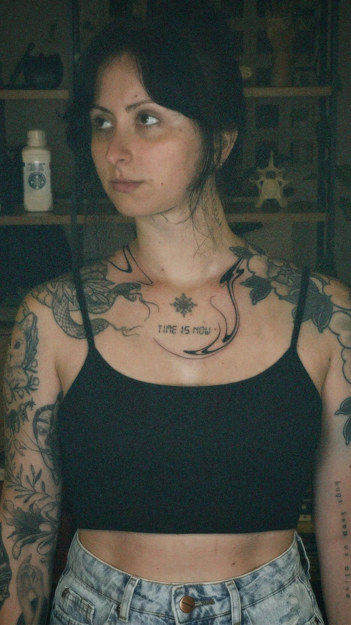 A young woman with black hair and tattoos on her arms and chest, wearing a black tank top and high-waisted jeans, standing indoors.