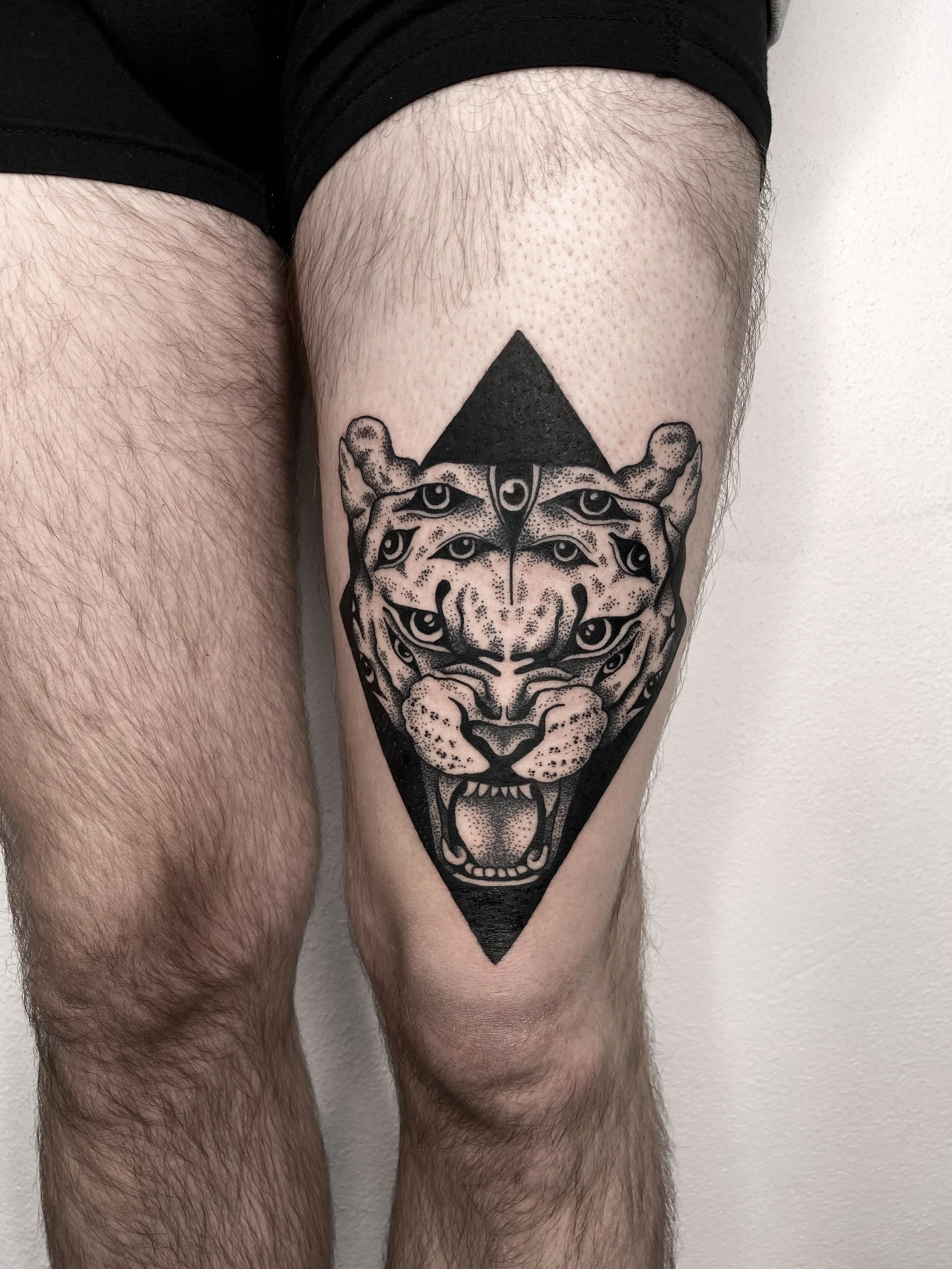 Tattoo of a tiger's face with multiple eyes inside a black diamond shape on a person's thigh.