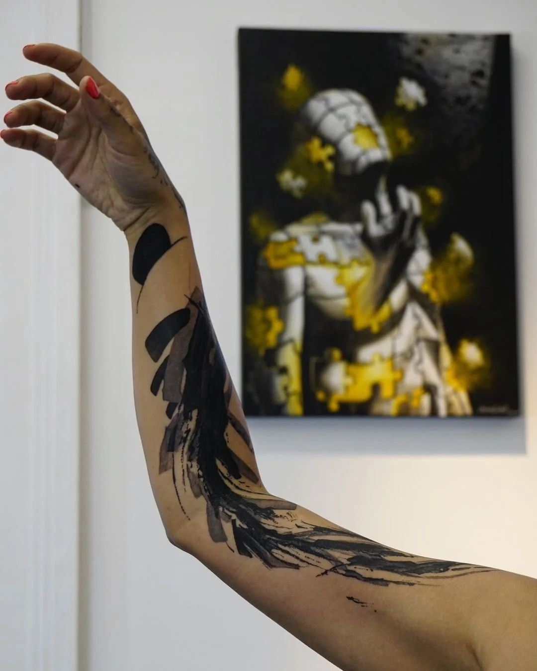 Abstract black ink arm tattoo with a background of a surreal artwork featuring a puzzle figure. The tattoo and painting share artistic themes, emphasizing creativity and imagination.