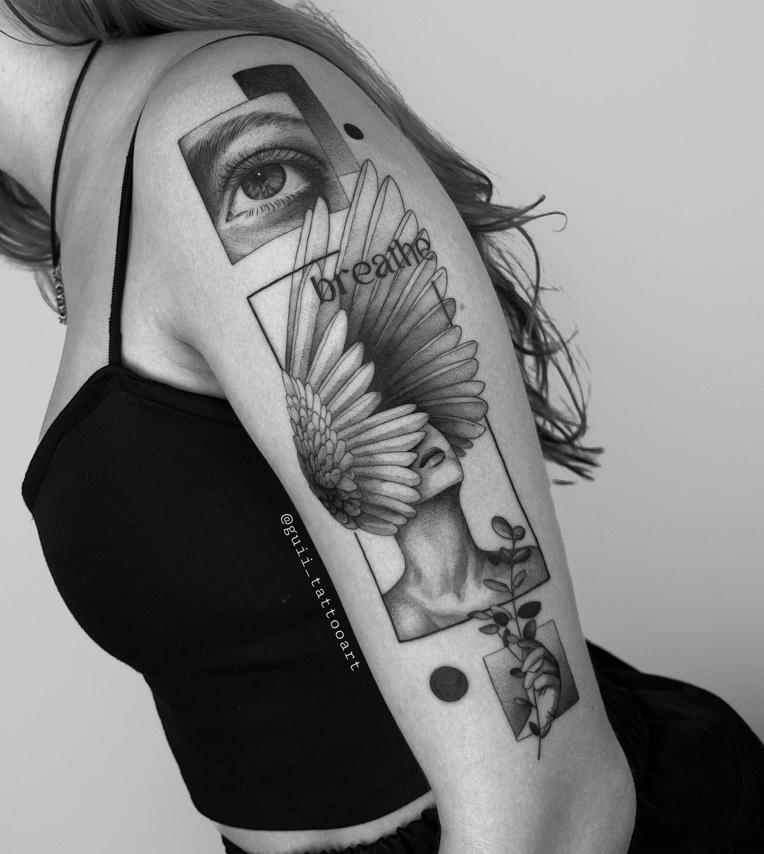Black and grey tattoo on a woman's upper arm featuring an eye, a woman's face with wings, geometric shapes, and the word 'breathing.'