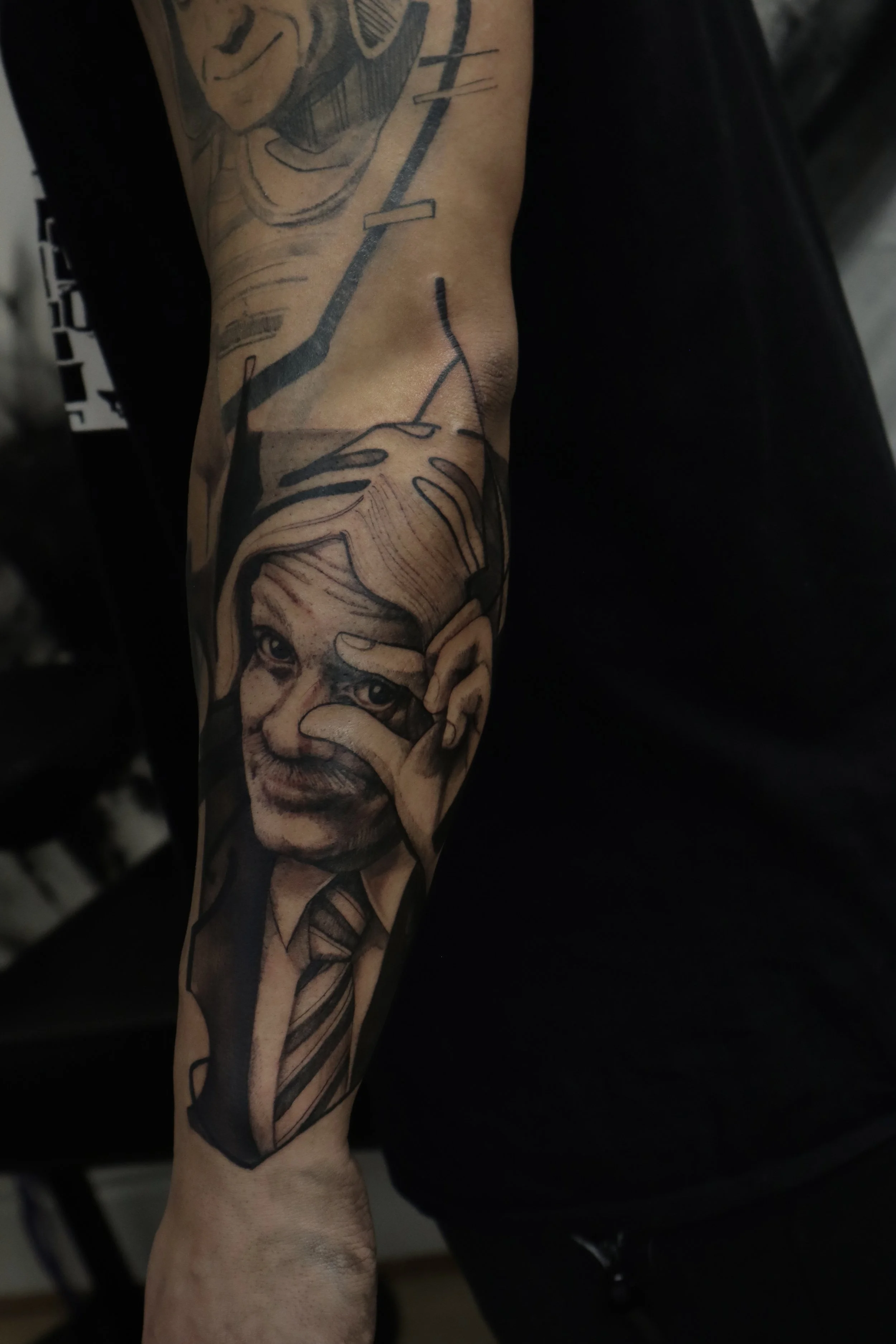 Black and gray tattoo on forearm of a man in a suit and tie, holding his hand near his eye like a lens.